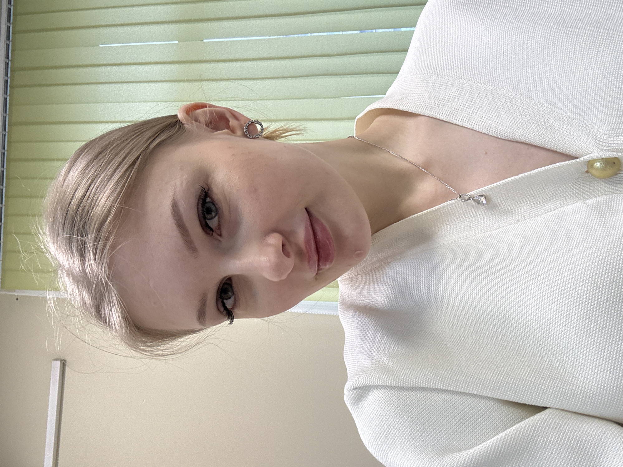 A young woman with blonde hair and fair skin is facing the camera. She is wearing a white textured top and a silver necklace with a pendant. She has a small, round earring in her left ear. The background consists of light yellow vertical blinds and a beige wall. The scene is possibly indoors. There is no visible information in the image to verify the location as Perm, Russia.