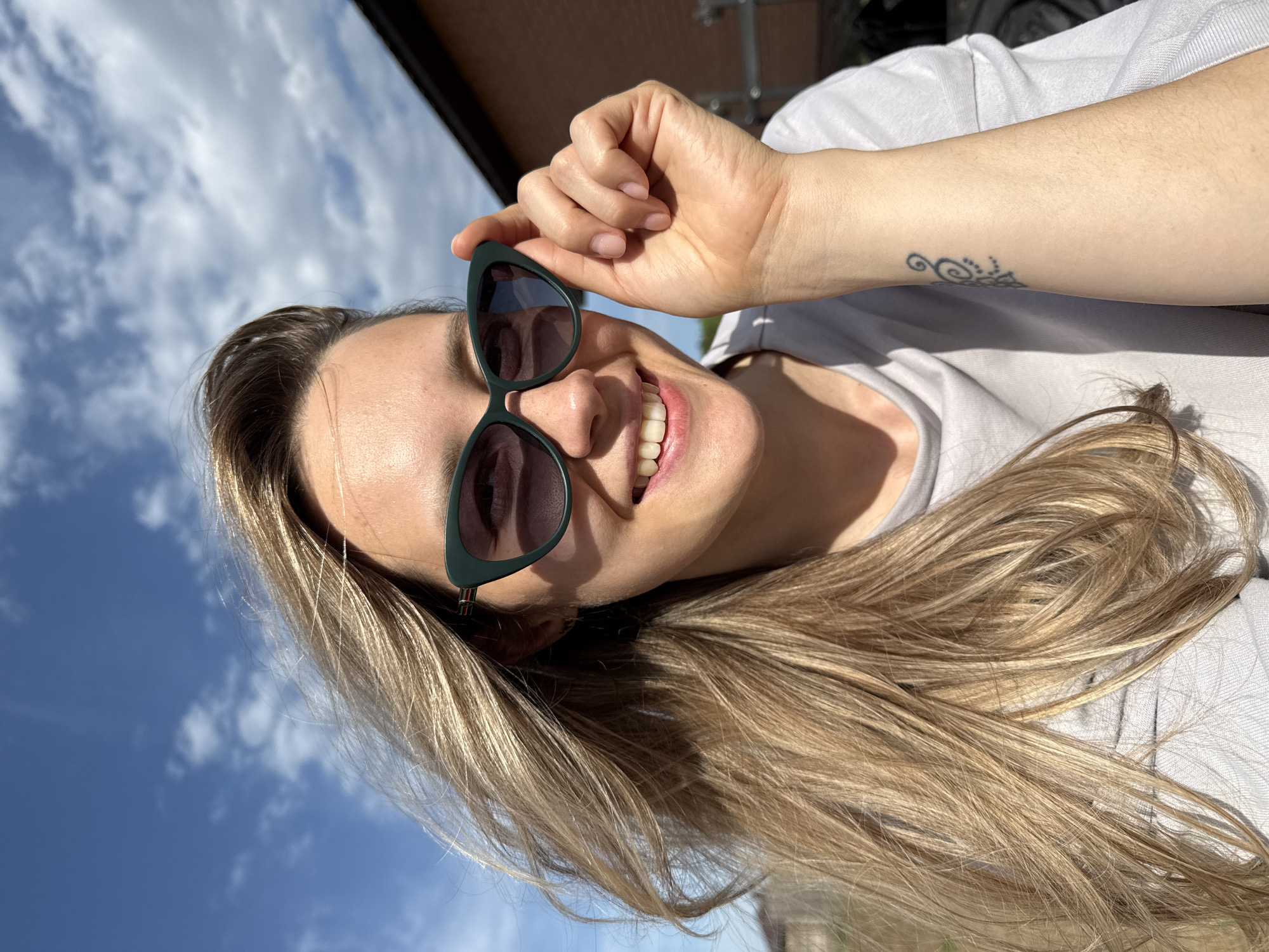 A medium shot depicts a smiling woman wearing sunglasses and a light gray t-shirt. She is holding the sunglasses with her right hand. The woman has long, blonde hair and a tattoo on her left wrist. The background shows a bright, blue sky with white clouds and a building partially visible in the upper left corner. The image appears to be taken outdoors in a sunny environment. There is no specific information about the location in Chaikovsky, Russia, provided in the image.