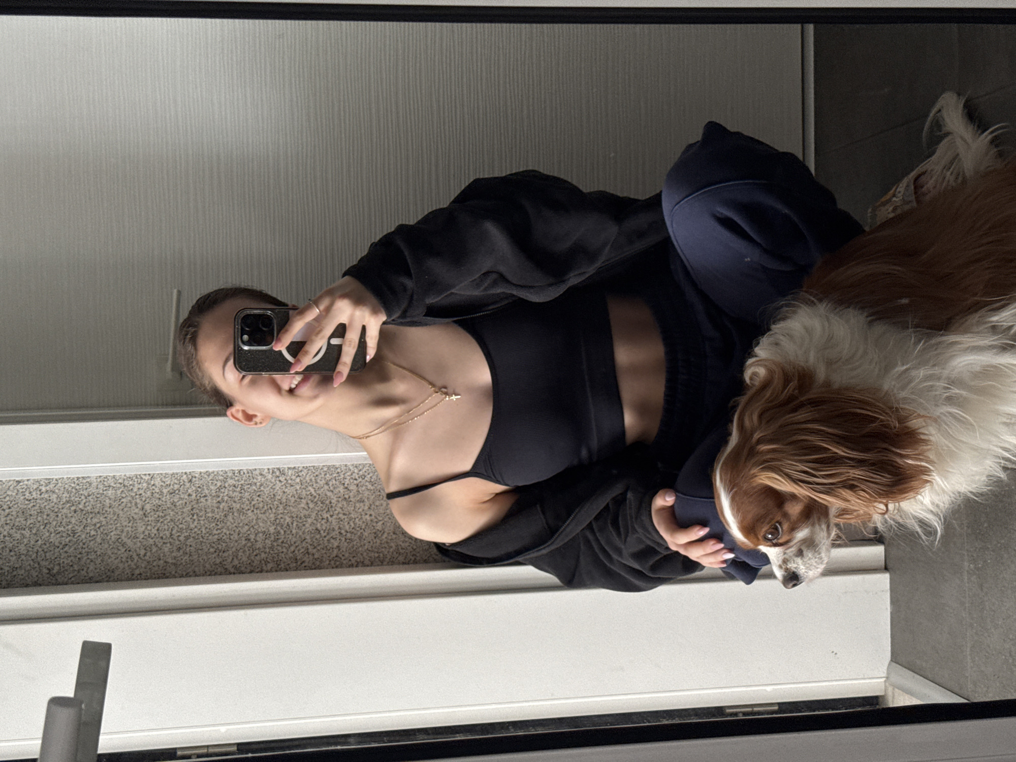 A young woman takes a selfie in a mirror. The woman is wearing a black sports bra, a black jacket, and navy blue sweatpants. She is holding a black smartphone up to her face with her right hand. She is smiling slightly. A Cavalier King Charles Spaniel dog is being held in her left arm. The reflection shows a hallway, with the floor, walls, and a door. The location is unknown.