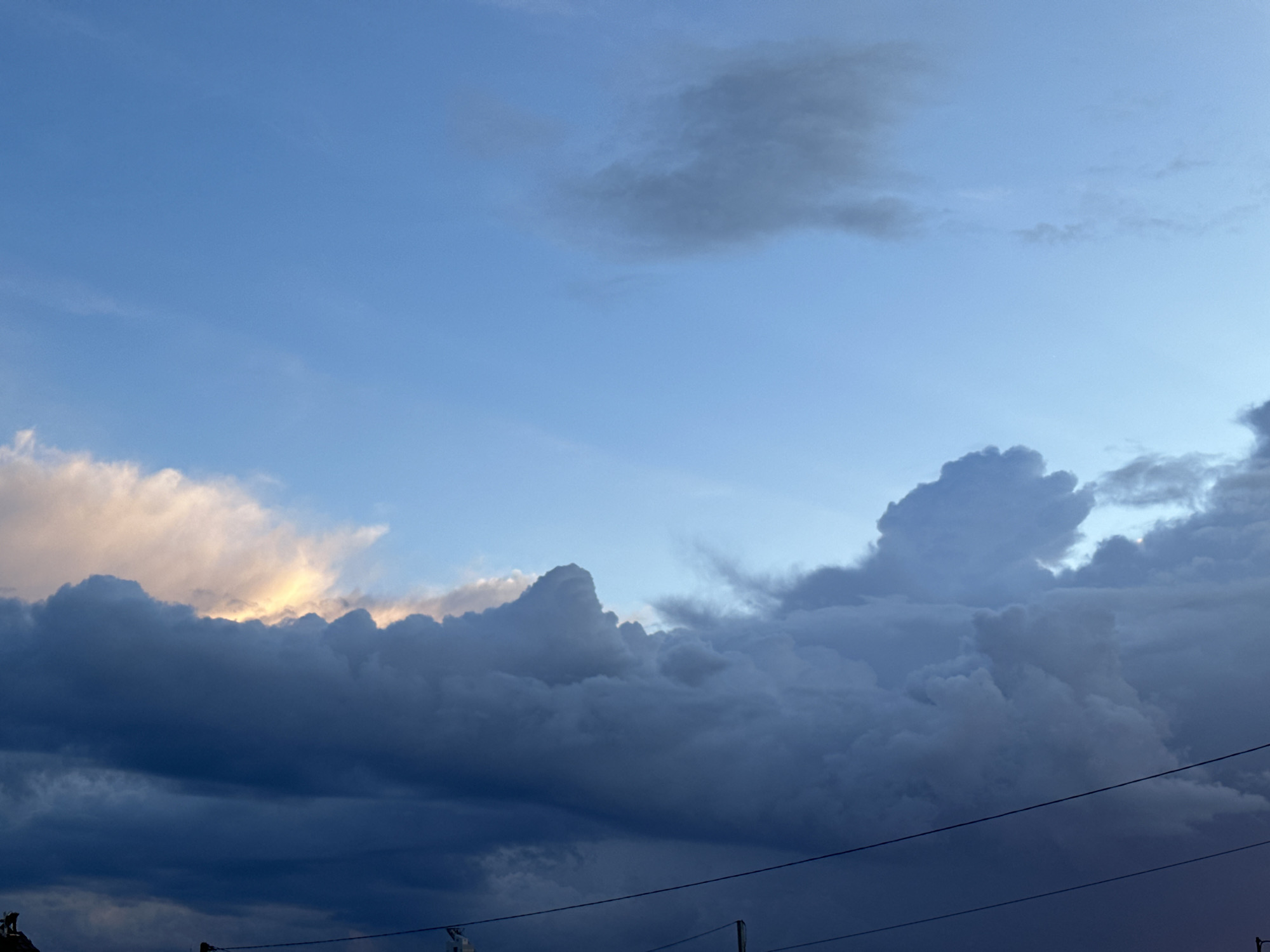The image shows a sky with a mix of blue and gray clouds. Some clouds are dark and looming, suggesting a storm, while others have a soft, golden light on their edges. There are power lines running across the bottom of the frame. At the very bottom left, there's a glimpse of a rooftop or building. The photo appears to be taken outdoors during the day. The location is Прикамский, Russia.