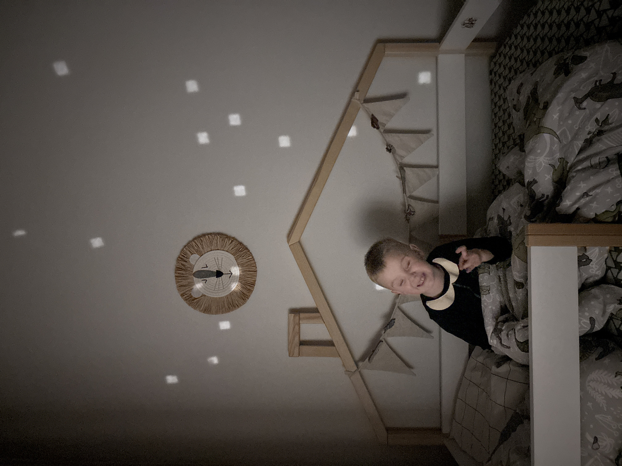 A low-angle, indoor shot reveals a child in a house-shaped bed. The bed is oriented vertically, with the headboard at the top of the image. The bed frame is wooden and painted white. A child with short blonde hair is lying in the bed, wearing a black long-sleeved shirt with a rounded collar. The child appears to be smiling. Bedding is a patterned gray and white. Above the bed is a ceiling with a circular straw-like decoration resembling a lion's face with a clock. The ceiling also shows multiple square-shaped light reflections. The scene is generally dimly lit. The image does not reveal the location as Chaikovsky, Russia.