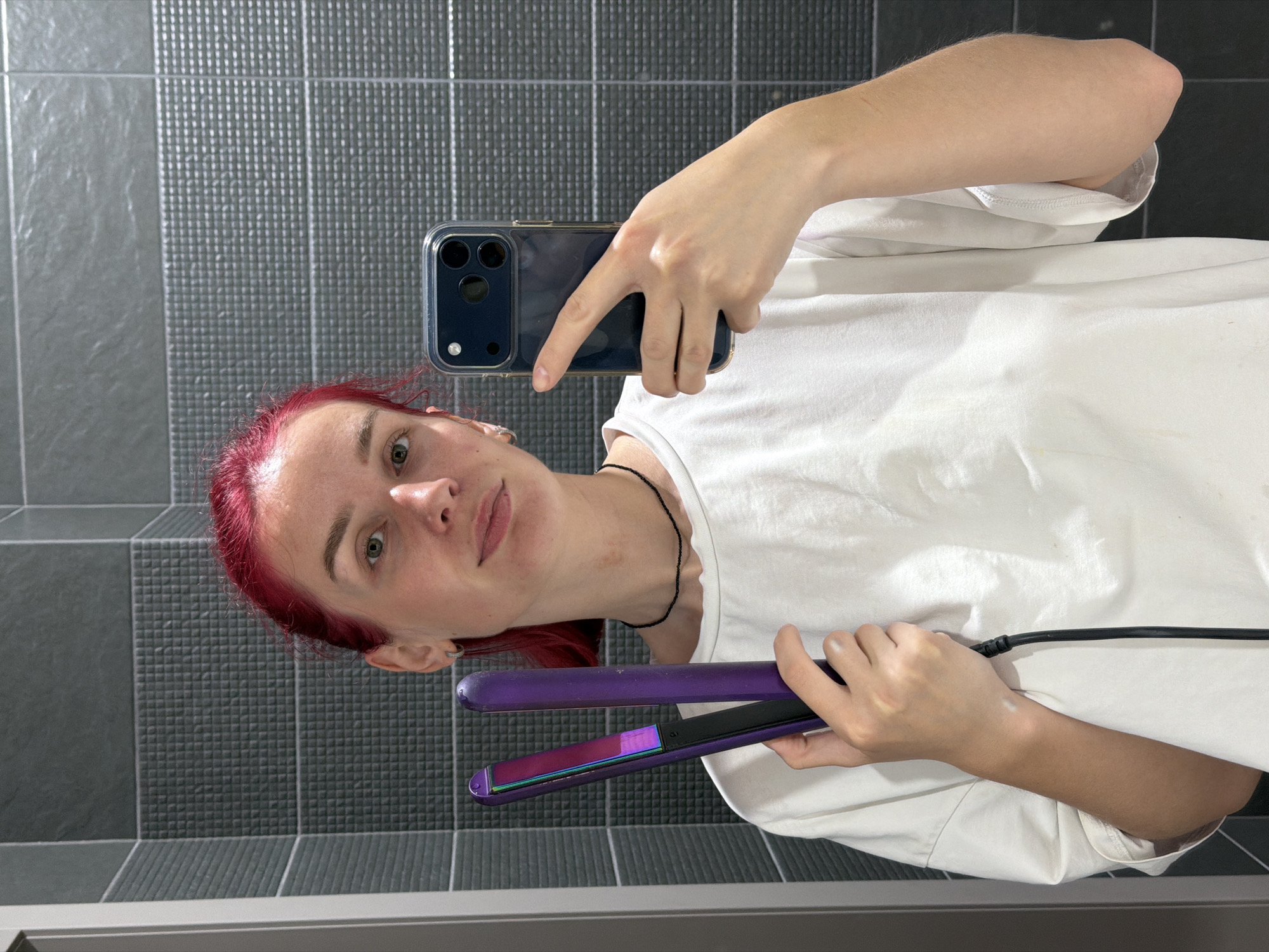 A person with bright red hair takes a mirror selfie while holding a purple hair straightener. The image is a reflection, showing the individual from the chest up. They are wearing a simple white t-shirt and have a light-colored necklace visible. Their right hand holds a dark-colored smartphone, likely an iPhone given the camera configuration, pointing it towards the mirror to capture the shot. In their left hand, they grasp a vibrant purple hair straightener with iridescent plates, connected to a black cord.
The person's expression is neutral to slightly smirking, with their eyes looking directly into the camera's reflection. Their face shows a few faint marks on the neck/collarbone area. The background behind the reflection consists of dark gray, rectangular tiles with a textured, small-grid pattern. Some tiles appear lighter or have a different finish, creating subtle horizontal banding. The lower edge of the image reveals a horizontal surface, possibly a vanity counter or bathtub edge, suggesting an indoor setting like a bathroom or dressing area in Chaikovsky, Russia. There is no visible text. The activity captured is a casual, self-portrait moment, possibly during a hair styling routine.