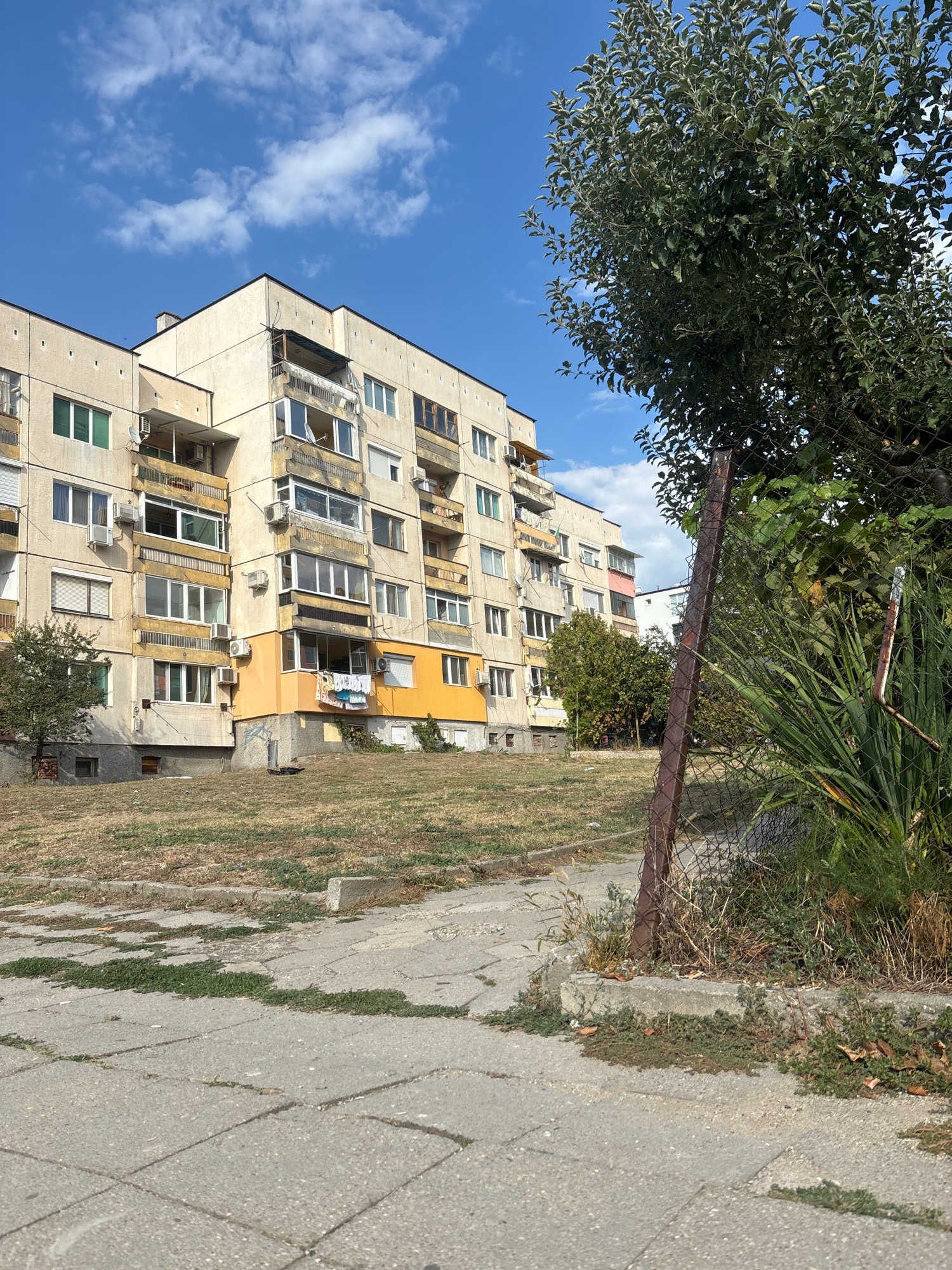 This media file shows an exterior view of a multi-story apartment building in Blagoevgrad, Bulgaria. The building appears to be constructed from concrete and has a beige exterior with some sections painted yellow and pink. Numerous windows and balconies are visible, some of which are enclosed and others have railings. Several air conditioning units are mounted on the exterior walls. There is laundry hanging on a clothesline on one of the balconies.
In the foreground, a paved pathway made of concrete slabs is visible, with patches of grass and weeds growing between the slabs and along the edge of the pathway. A slope covered in dry grass leads up to the building. A chain-link fence with a rusty metal post runs along the right side of the frame, partially obscuring a dense area of green foliage and plants. A large tree with green leaves is situated in the upper right corner, casting a shadow. The sky is blue with scattered white clouds, suggesting a clear day. The overall impression is that of an urban residential area, possibly in a slightly weathered state. The time of day appears to be daytime, and the weather is sunny. There is no visible text in the image.