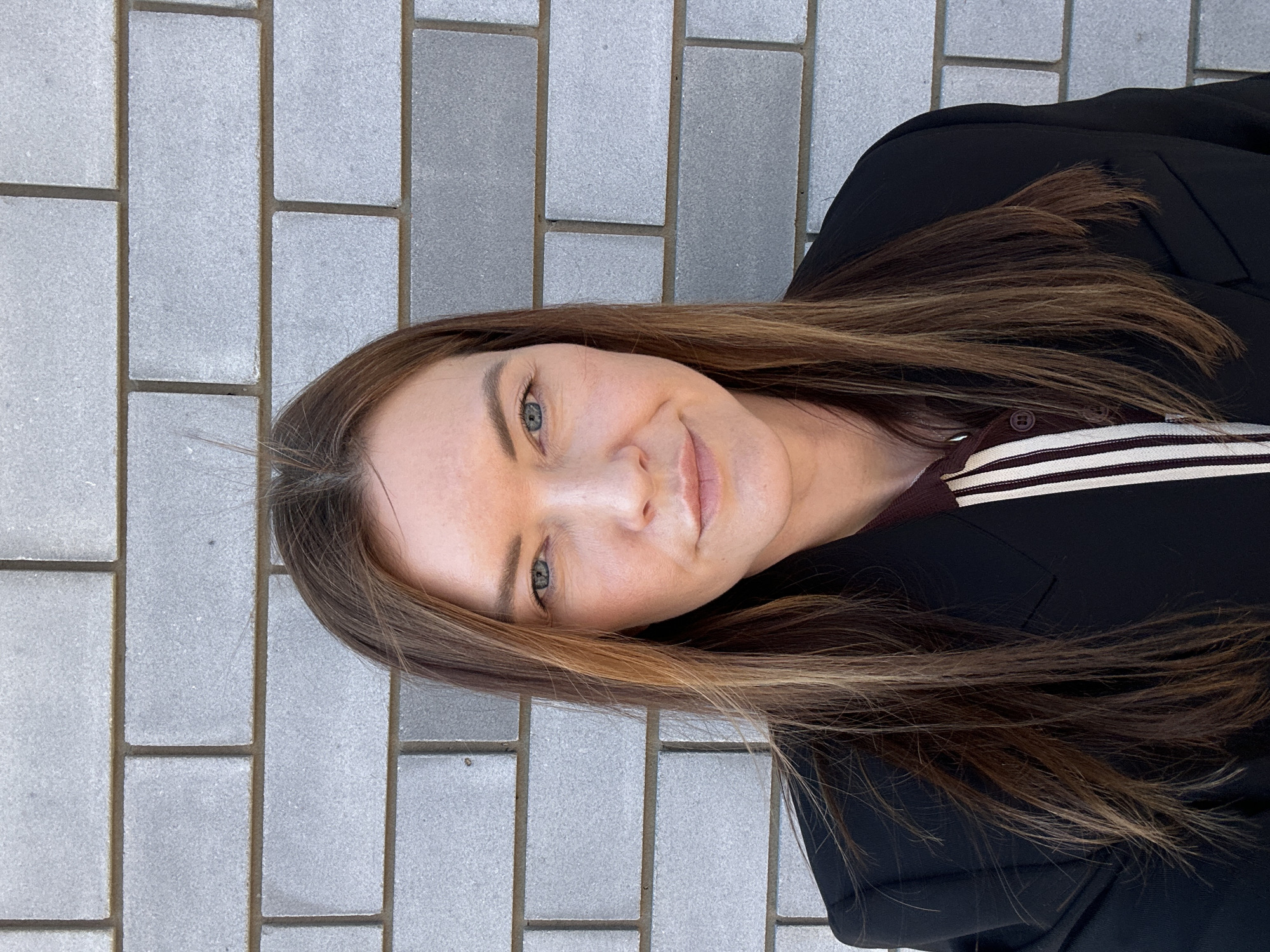 A medium, slightly off-center shot depicts a woman with long, light brown hair that is subtly highlighted. Her hair cascades down her right shoulder. The woman has light skin, blue eyes, and a gentle smile. She is wearing a black blazer over a top with burgundy and white horizontal stripes. The background features a pavement of rectangular gray bricks. The location is Прикамский, Russia.