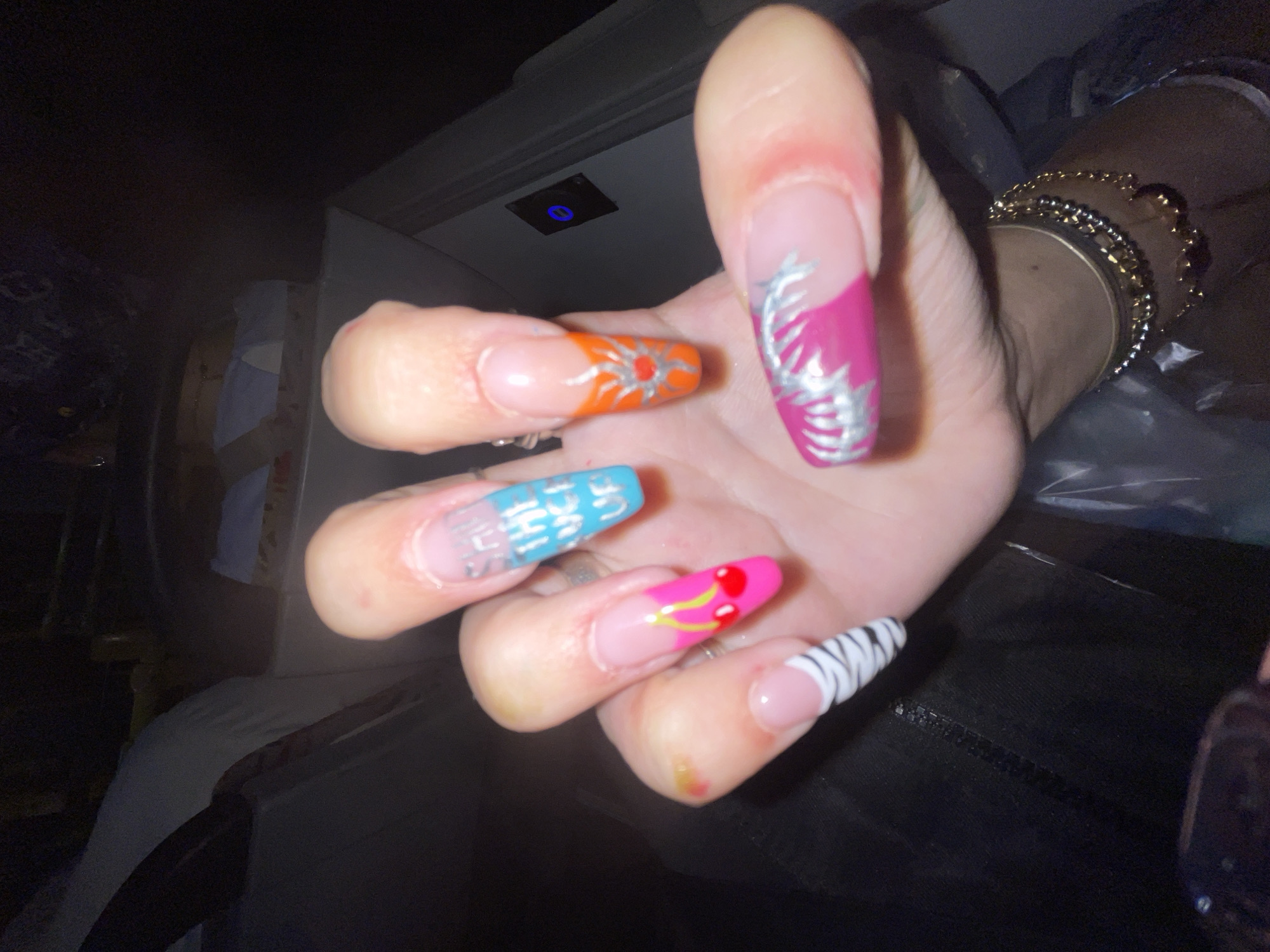 A close-up shot shows a person's hand with meticulously designed artificial nails. The nails feature various vibrant designs, including a pink background with cherry art on one finger, a blue and white gradient with text on another, a sunburst design in orange and red, a pink and silver palm tree design, and a black and white zebra print. The hand also displays some minor skin abrasions and is adorned with multiple gold and silver necklaces. The background is dark and out of focus, suggesting an indoor setting with artificial lighting. The context of Raanana, Israel, is not visually evident in the image itself.