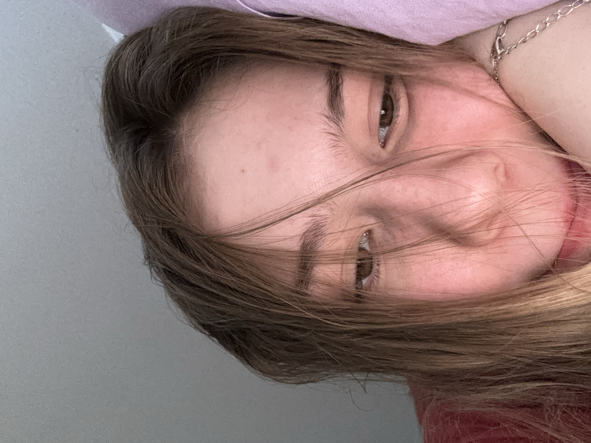 The image is a close-up, low-angle portrait of a young person with long, light brown hair. Their face is partially obscured by their hair, and they are resting their head on a pink pillow or blanket. The person's eyes are looking downwards. They are wearing a red top. The background is a plain gray wall and a pink textile. The image was taken in Sinyavino, Russia.