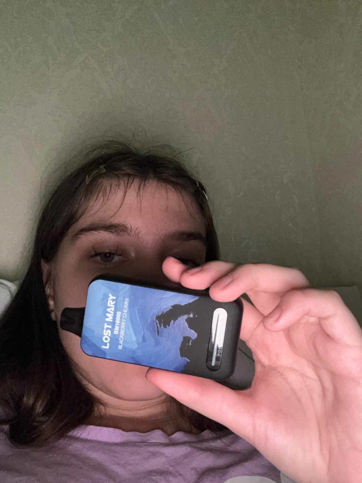 A young person is holding a black and blue electronic cigarette, partially obscuring their face. The cigarette has text that reads "LOST MARY" and "BLACKBERRY CHERRY". The background is a textured, light green wall. The person is wearing a purple shirt. The image appears to be taken indoors. There is no visual information in the image that would indicate the location is in Zavolzhye, Russia.