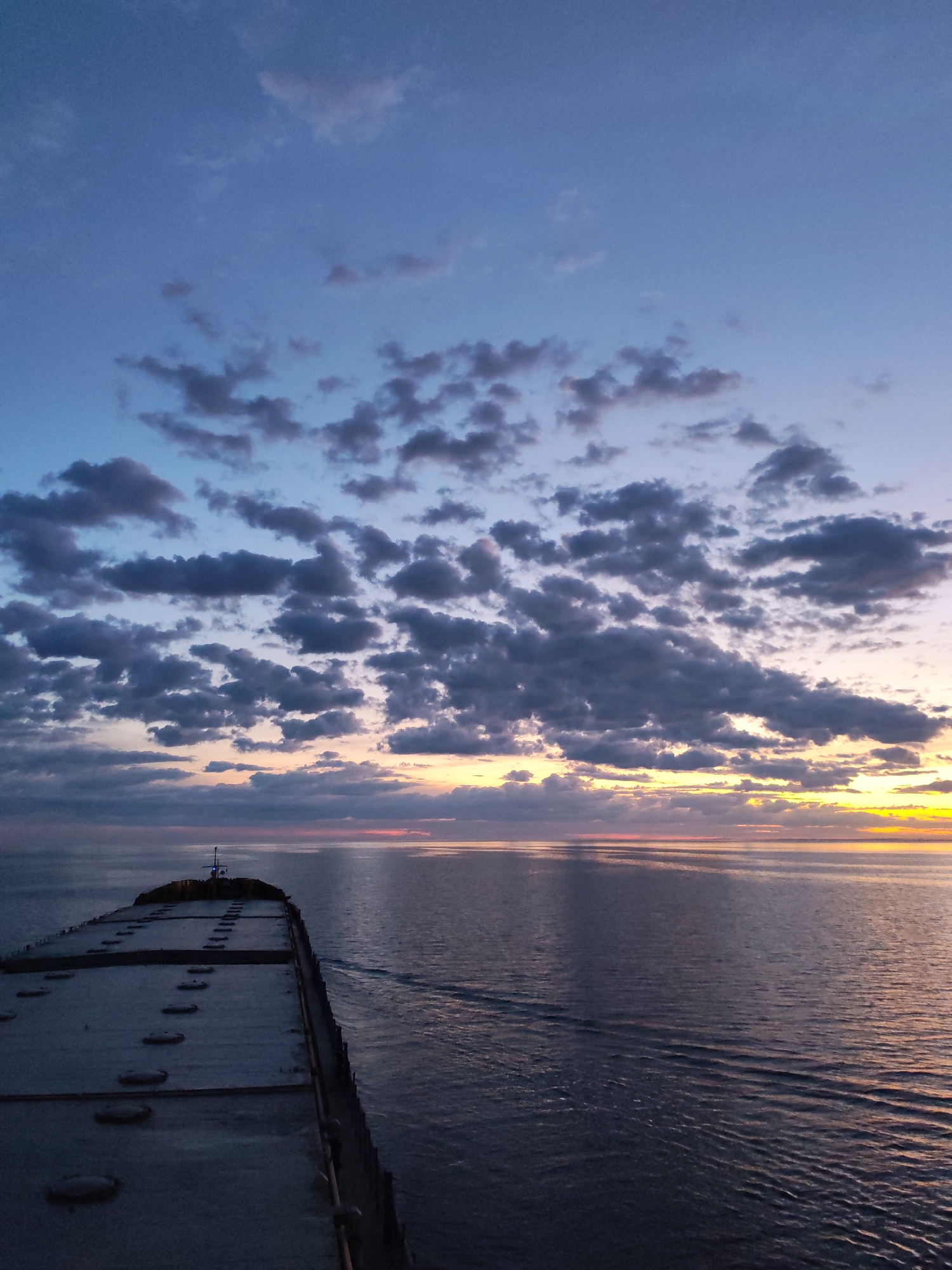 The image depicts a sunset over the ocean from the deck of a ship. The sky is filled with clouds, illuminated by the setting sun. The ship is moving, as evidenced by the wake in the water. The location is unknown.