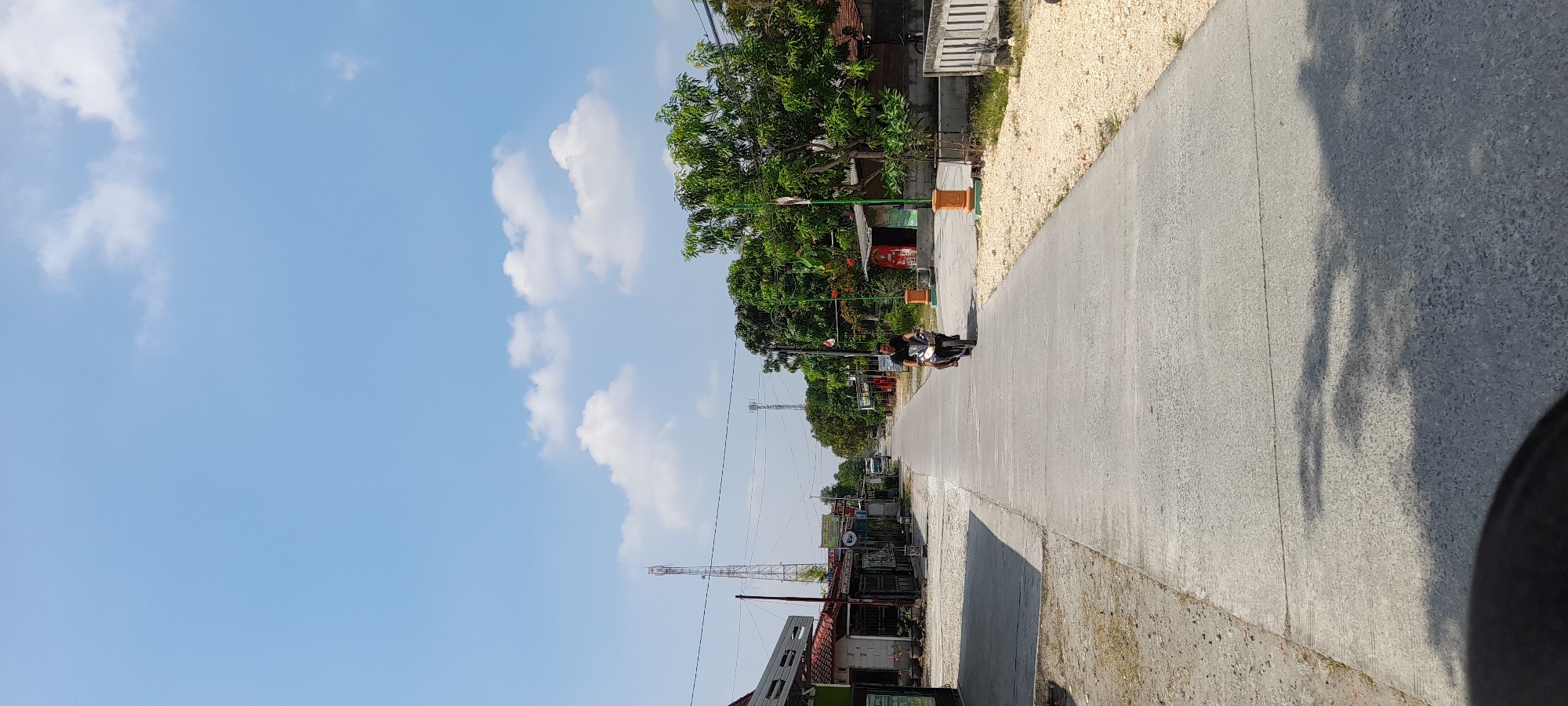 The image depicts a sunny day in Kuwu, Indonesia, showing a concrete road in a rural or suburban area. A person is riding a motorcycle down the center of the road, facing away from the camera. The road is flanked by buildings on both sides, with some trees and vegetation visible. In the distance, there are communication towers. The sky is blue with scattered white clouds. The ground on the sides of the road is covered with gravel and dirt. The scene appears peaceful and ordinary.