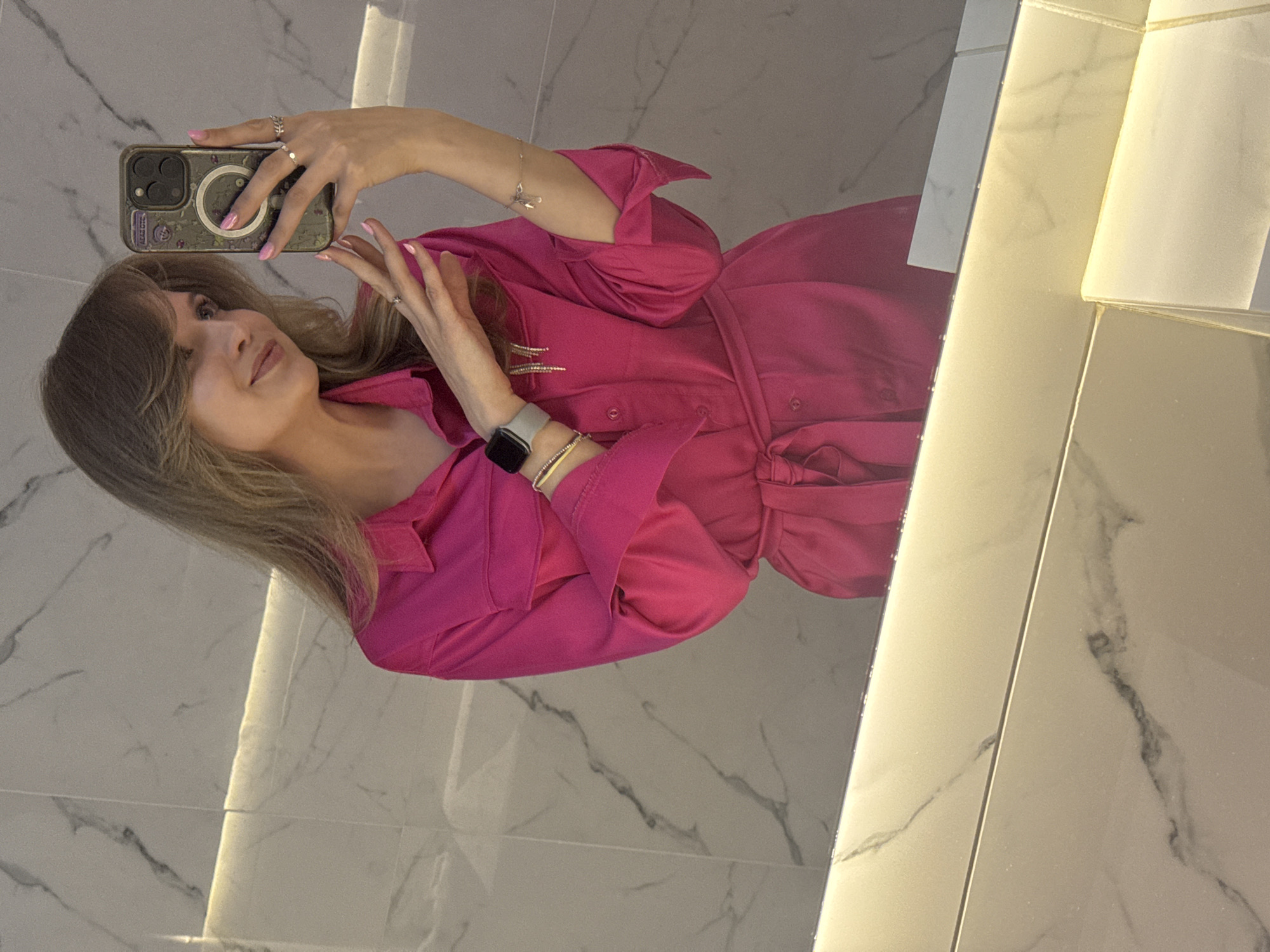 A woman is taking a selfie in a bathroom. She is wearing a pink dress with sleeves and a belt around her waist. She has long, blonde hair with bangs. She is holding a smartphone in her left hand and posing with her right hand. She has a smartwatch and bracelets on her left wrist, and rings on her fingers. The background is a bathroom with marble walls. The location of the bathroom in the city of Izhevsk, Russia is unknown.