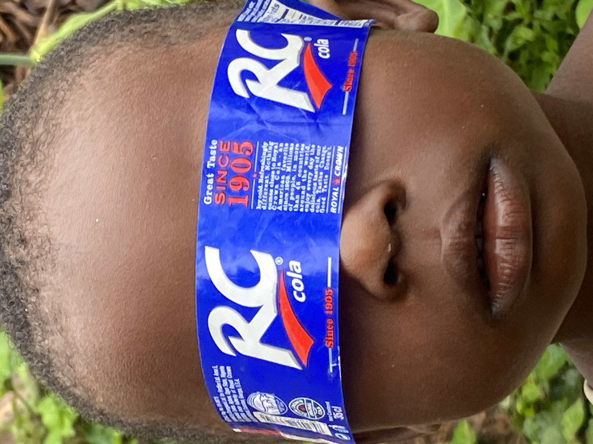 The image is a close-up, eye-level shot of a young child's face, centered vertically and horizontally. The child's eyes are covered by a blue and white paper band, which appears to be a label from an RC Cola product. The band has the "RC cola" logo prominently displayed, along with text such as "Great Taste Since 1905" and "Royal Crown". The child's dark skin tone and the texture of their hair are clearly visible. The background is softly blurred and consists of green foliage, suggesting an outdoor setting, possibly in a rural or natural environment. The image composition focuses tightly on the child's face, drawing attention to the unusual placement of the RC Cola band over their eyes. There is no specific indication of the city of Ufu Esioni, Nigeria within the image itself.