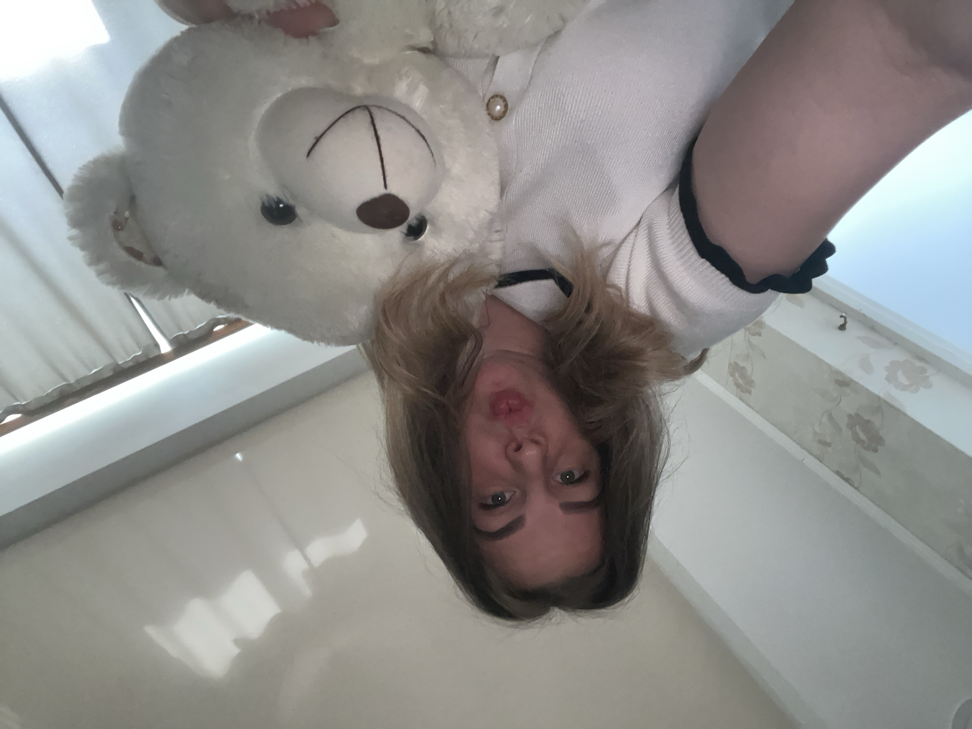 A woman with shoulder-length brown hair is holding a white teddy bear. She is wearing a white knitted sweater with a black trim. The photograph is taken from a high angle, looking down at the woman and teddy bear. The background includes a white wall and window. There is no information to suggest that this image was taken in Izhevsk, Russia.