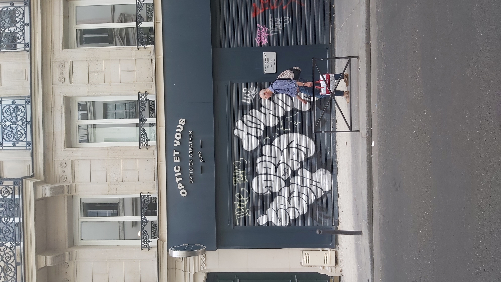A middle-aged man with glasses walks past a store with a closed metal security shutter on a street in Paris, France. The store is aoptician's shop named "OPTIC ET VOUS - OPTICIEN CREATEUR - paris". The shutter is covered in large white graffiti. The man is wearing a blue and white patterned shirt, dark pants, and brown shoes. He is carrying a backpack and a red and white shopping bag. The street is paved with asphalt and there is a sidewalk in front of the store. The buildings in the background are made of light-colored stone. The weather appears to be overcast.