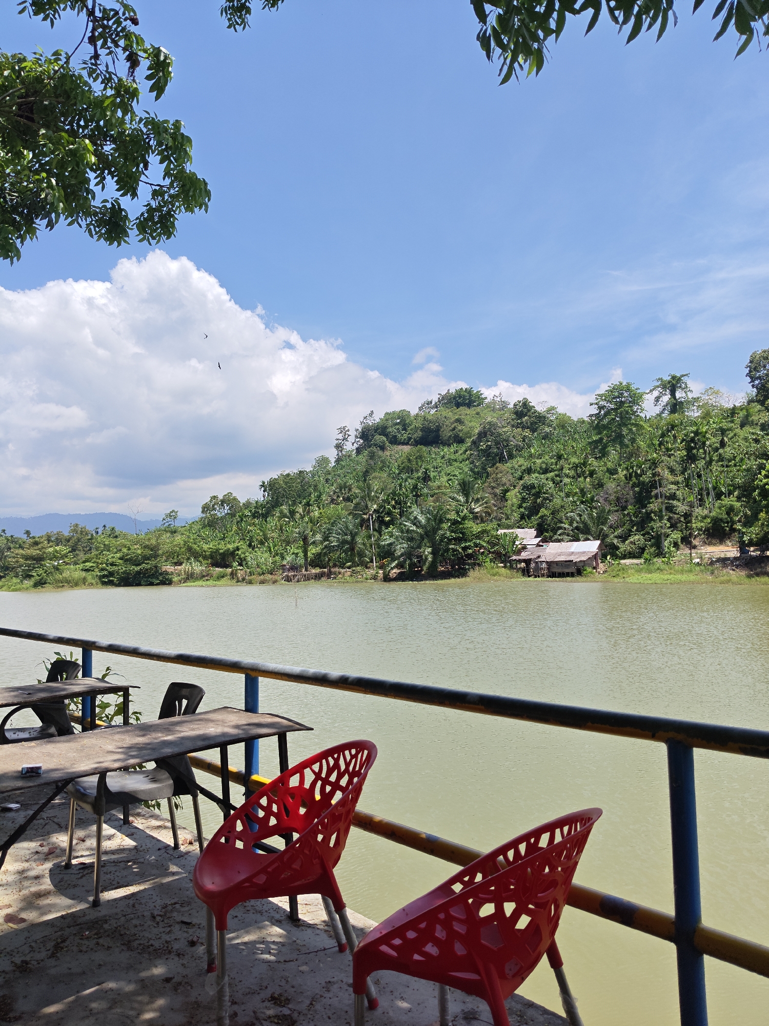 A daytime outdoor scene features a lake and lush vegetation. A railing with blue, yellow, and black paint runs horizontally across the lower portion of the frame. In front of the railing, a set of weathered tables and chairs are arranged, including two bright red plastic chairs. The lake is placid, with a murky, greenish-brown color. On the opposite shore of the lake, a hillside is covered in dense green trees and vegetation, with a small, dilapidated structure visible among the trees. The sky is blue with scattered white clouds. Tree branches and leaves are visible in the upper left and right corners of the image.