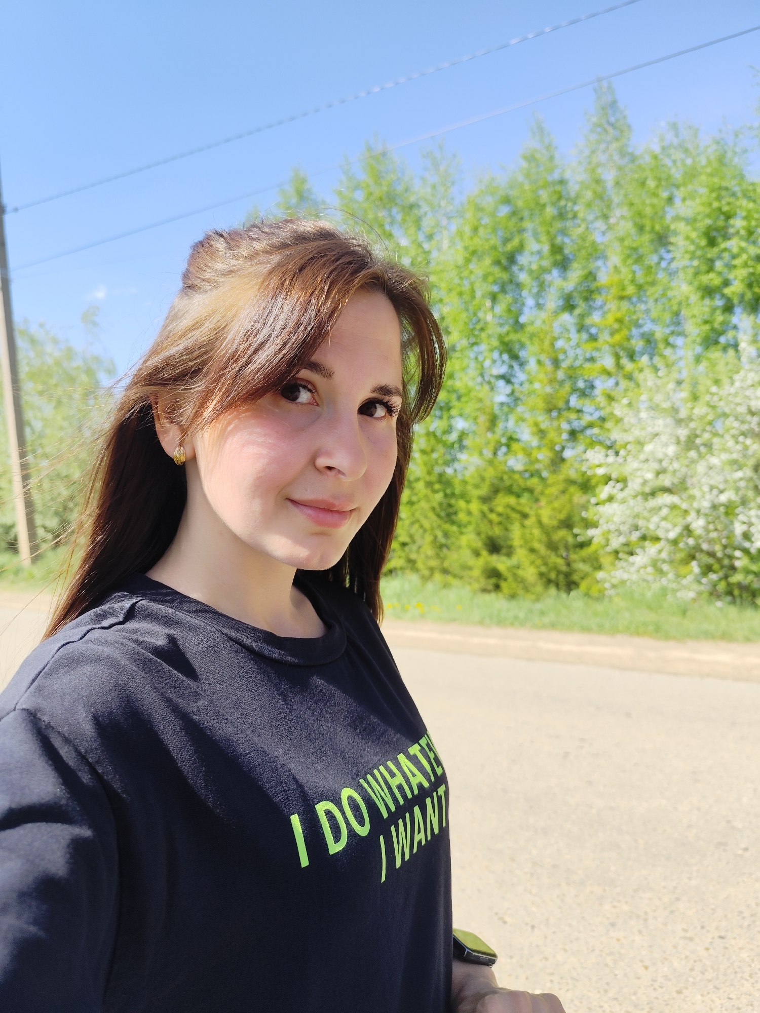A woman with long brown hair stands outside in Фоки, Russia. She wears a black T-shirt with the words "I DO WHATE I WANT" in neon green letters. The background consists of trees and a blue sky.