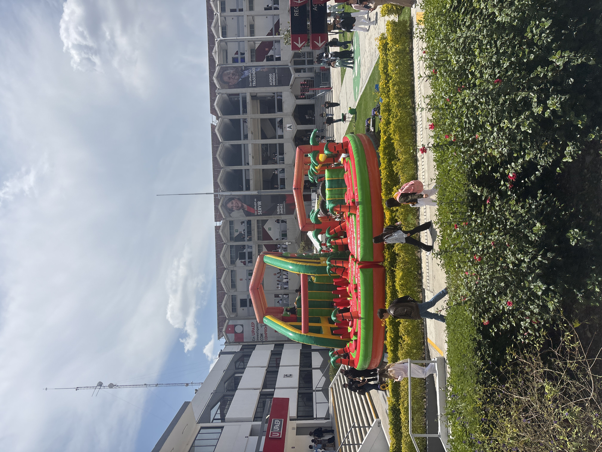 The image shows an outdoor scene in Puebla, Mexico. An inflatable obstacle course with orange, green, and red stripes sits on a paved area, surrounded by low hedges and a bush with red flowers. People are walking by the obstacle course. Behind the inflatable structure is a multi-story building with a modern design. The building has hexagon-shaped window structures, and posters with the names Fabila Jimenez, Alfonso Mendoza and the UPAEP logo are visible. The sky is cloudy with a blue hue. A set of stairs with a metal railing leads up to the paved area and the obstacle course.