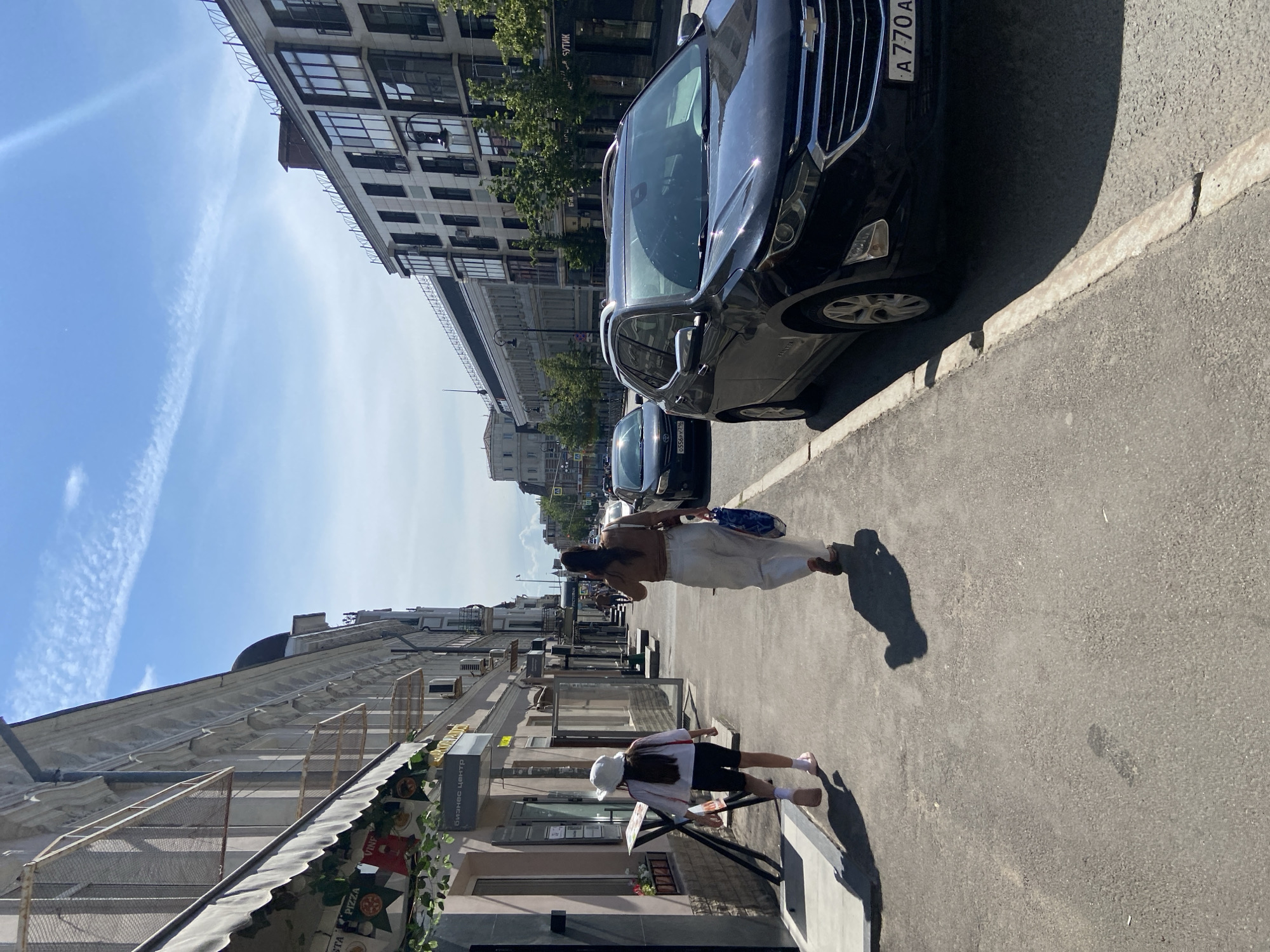 This detailed image depicts a sunny urban street scene in Kazan, Russia, during what appears to be late morning or early afternoon.
The primary focus is a paved asphalt sidewalk on the left, where two females are walking away from the camera. The closer individual is a young female, possibly a child or teenager, wearing a white wide-brimmed hat, a white long-sleeved top with red trim, black cycling shorts, and pink slide sandals. Her right sock is bunched around her ankle. Behind her walks an adult female with long dark hair, wearing a brown crop top, loose-fitting white trousers, and dark flat shoes. She carries a blue and white patterned reusable bag in her left hand. Both individuals cast long, distinct shadows to their right, indicative of strong overhead sunlight.
To the left of the sidewalk, a multi-story building with a light-colored facade (likely white or cream) features ornate cornices and multiple windows. A grey awning extends over a ground-floor storefront. Visible signage includes a "Pizza" graphic and a sign with Cyrillic text "Бизнес центр" (Business Center). Metal mesh panels are attached to the upper windows, and several external air conditioning units are mounted on the wall.
To the right of the sidewalk is a street with parked vehicles. In the foreground, a black Chevrolet Equinox SUV is parked parallel to the curb. Its front license plate displays "A 770 A." Further down the street, another dark-colored sedan is parked, with a partially visible license plate "O 556 P Y 16," where "16" is the regional code for Tatarstan, confirming the location as Kazan. Beyond the parked cars, the street extends into the distance, with additional vehicles and pedestrians.
The buildings on the right side of the street are more modern in appearance, featuring multiple stories with extensive glass windows or balconies and a darker facade. Small trees are planted at intervals along the sidewalk fronting these buildings. A sign with the Cyrillic word "БУТИК" (Boutique) is partially visible on one of these buildings.
The sky overhead is clear and bright blue, with scattered wispy white clouds and a faint contrail or cloud streak visible in the upper right quadrant. The overall impression is a bustling urban environment on a pleasant day.