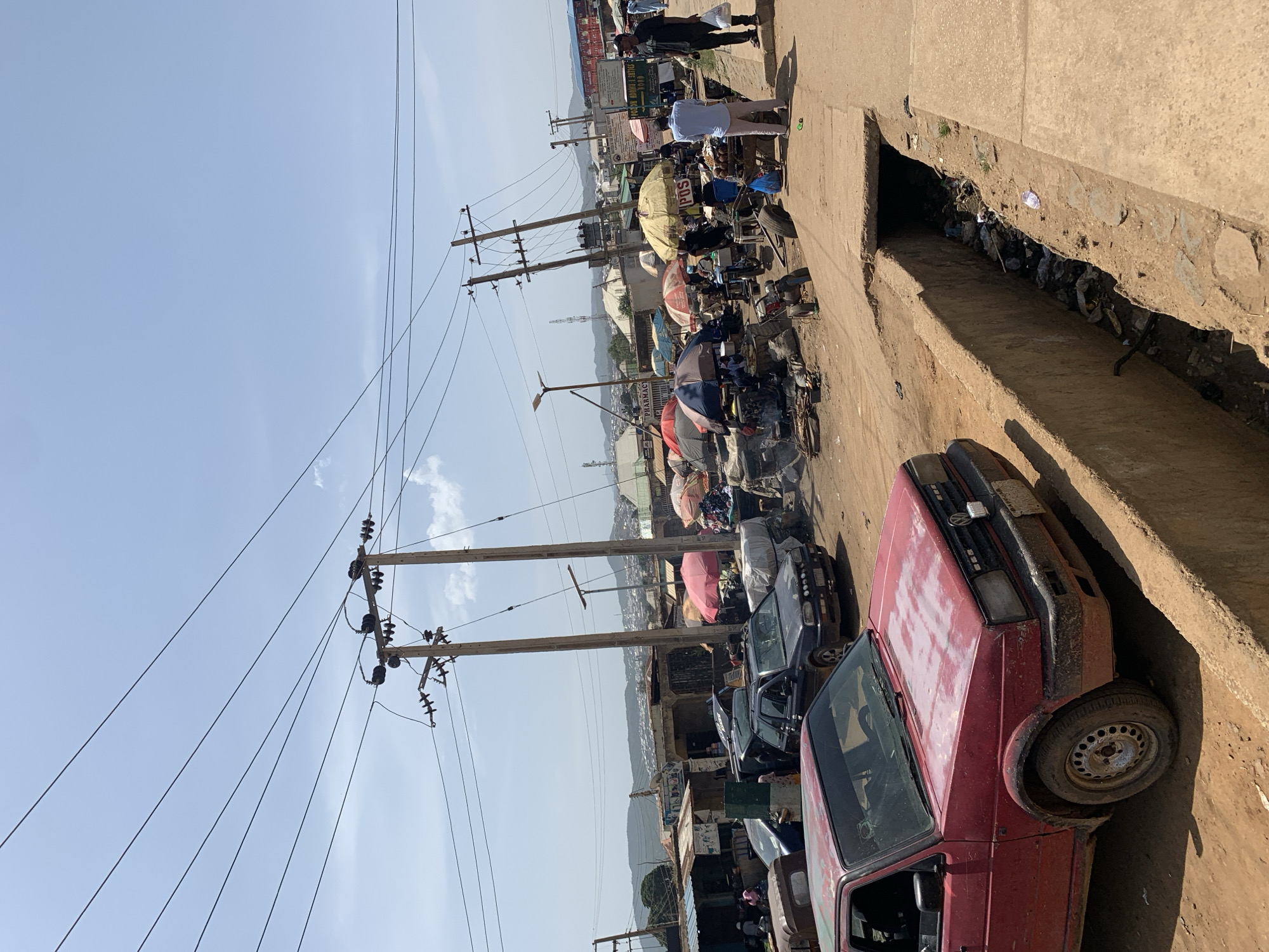 The image shows a street scene in Rafin Par, Nigeria. The street is lined with parked cars and street vendors with umbrellas and stalls. Overhead power lines stretch across the sky. A drainage ditch runs alongside the road, partially filled with debris. People are walking along the sidewalk. A red car is parked prominently in the foreground. The background features a cityscape with buildings and a distant mountain range.