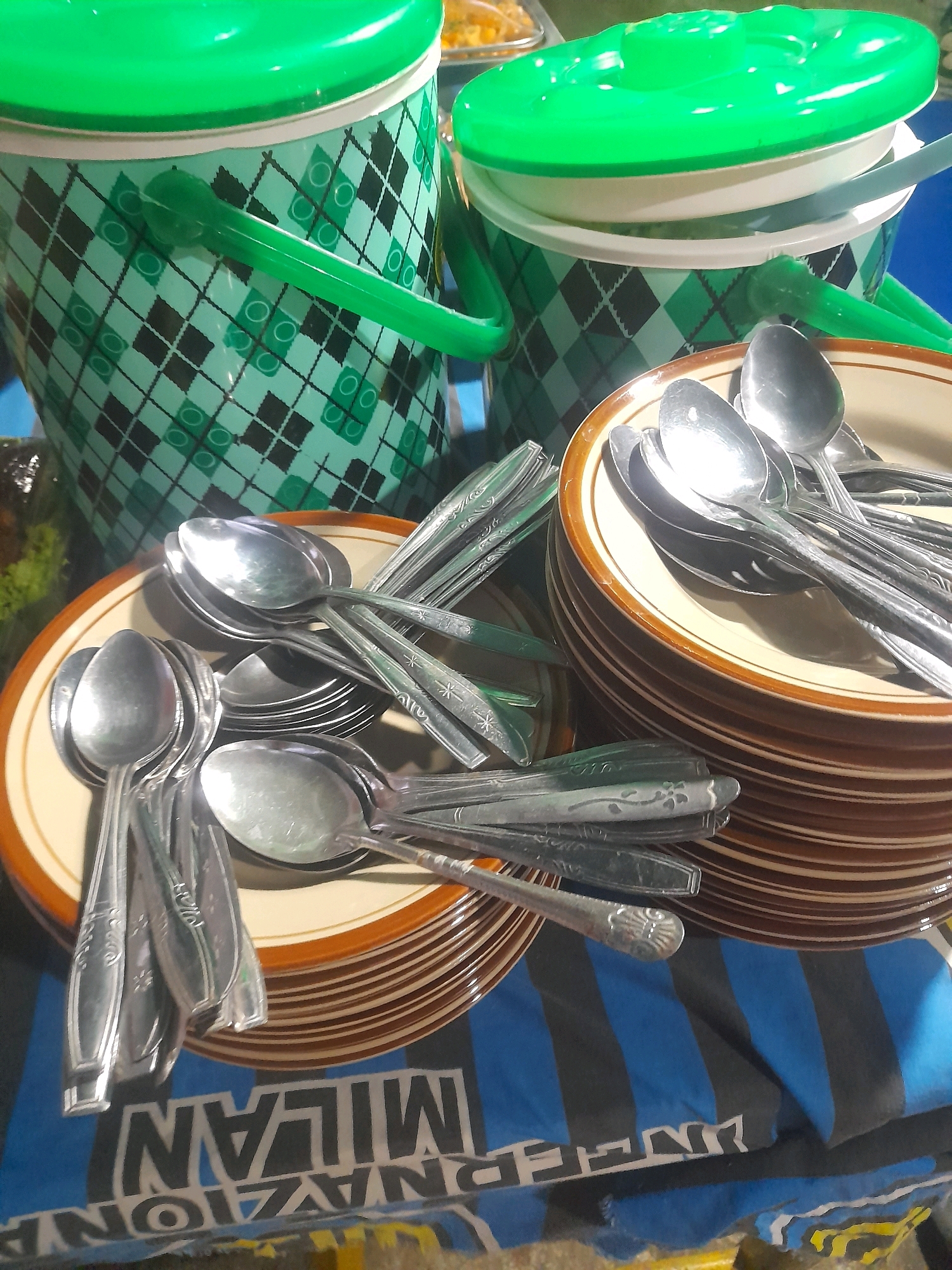 In the city of Mlaras, Indonesia, this image depicts a collection of metal spoons stacked on top of brown and white plates, which are then placed on a blue and black striped surface that appears to be a table. In the background, there are two green buckets with diamond patterns and green lids. A partially visible dish of food is also present behind the buckets. The overall scene suggests a gathering or a meal being prepared or served.