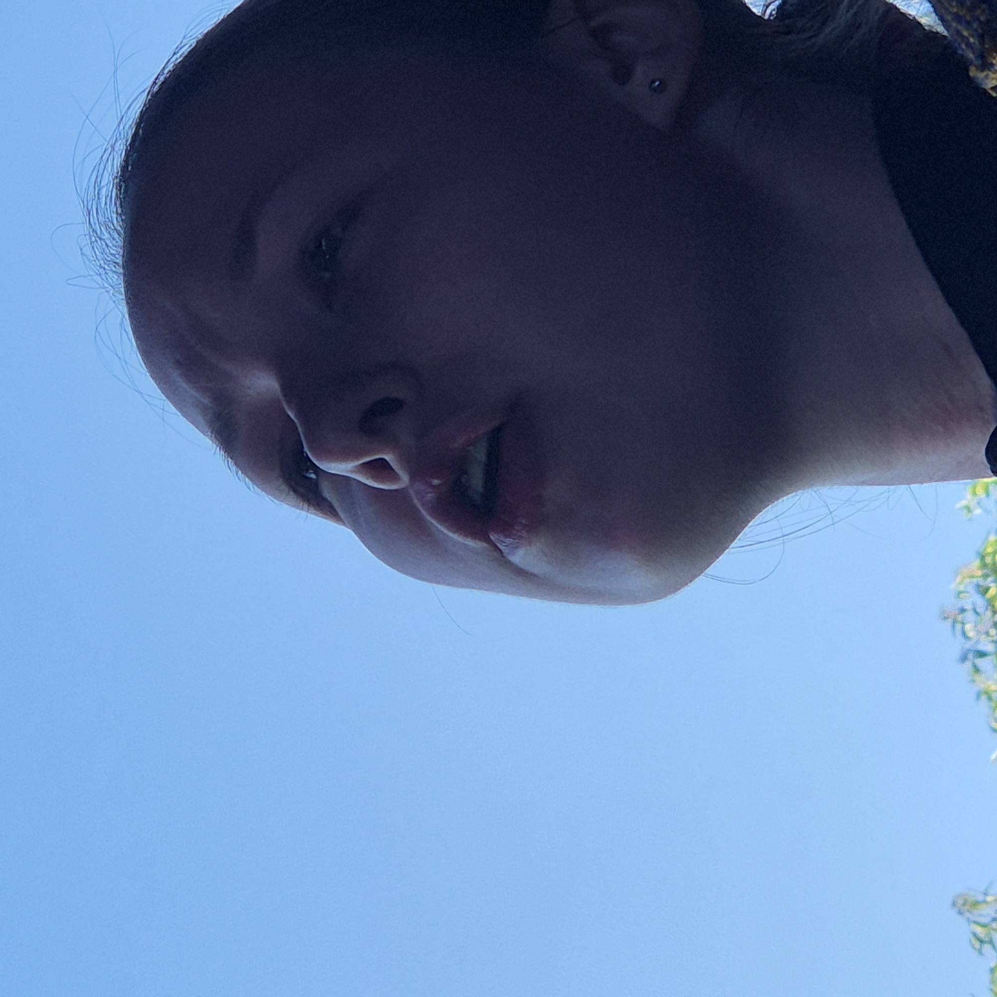 The image shows a close-up, low-angle shot of a young person's face looking upwards against a bright blue sky. The person's face is partially obscured by shadow, but their mouth is slightly open, and their brow is furrowed. Some wisps of hair are visible against the sky. The lower part of the image shows a blurred hint of green foliage. The scene appears to be outdoors on a clear day. This image was taken in Novo-Stroika, Russia.