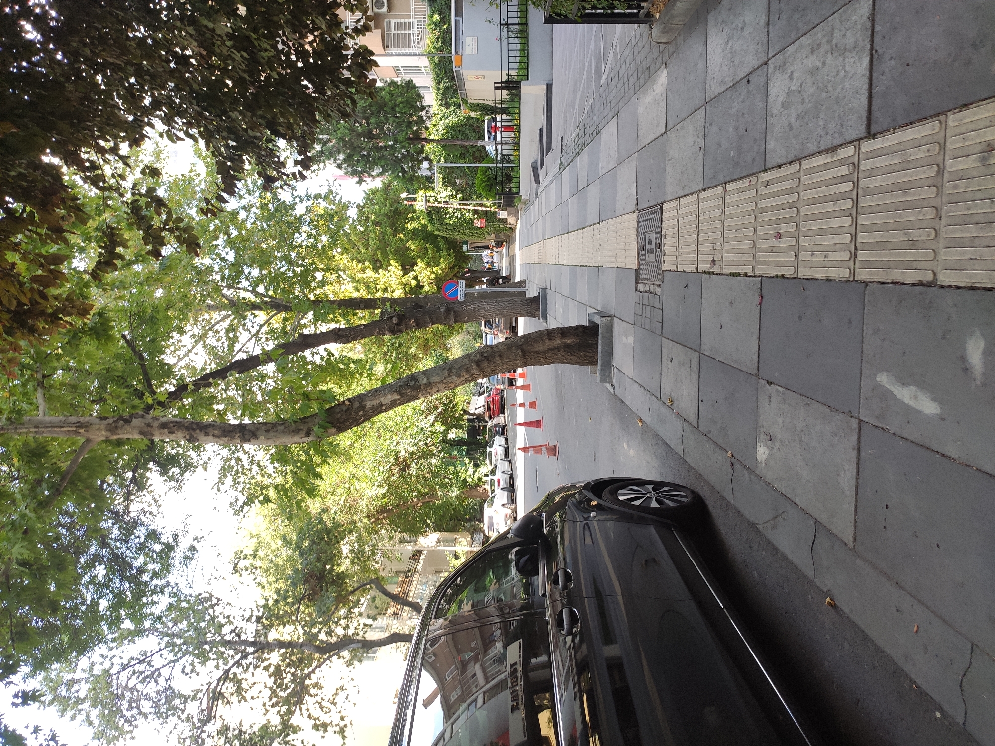 This is a street scene in Ankara, Turkey, with a focus on a sidewalk and adjacent street. A black van is parked on the left side of the image, partially obstructing the view of the street which has several cars parked and in motion, as well as orange traffic cones. The sidewalk is paved with large grey tiles and features a tactile paving strip for visually impaired pedestrians. Trees line both sides of the street, casting dappled shade. Buildings are visible in the background, suggesting an urban environment. The sky is bright, indicating it is daytime.