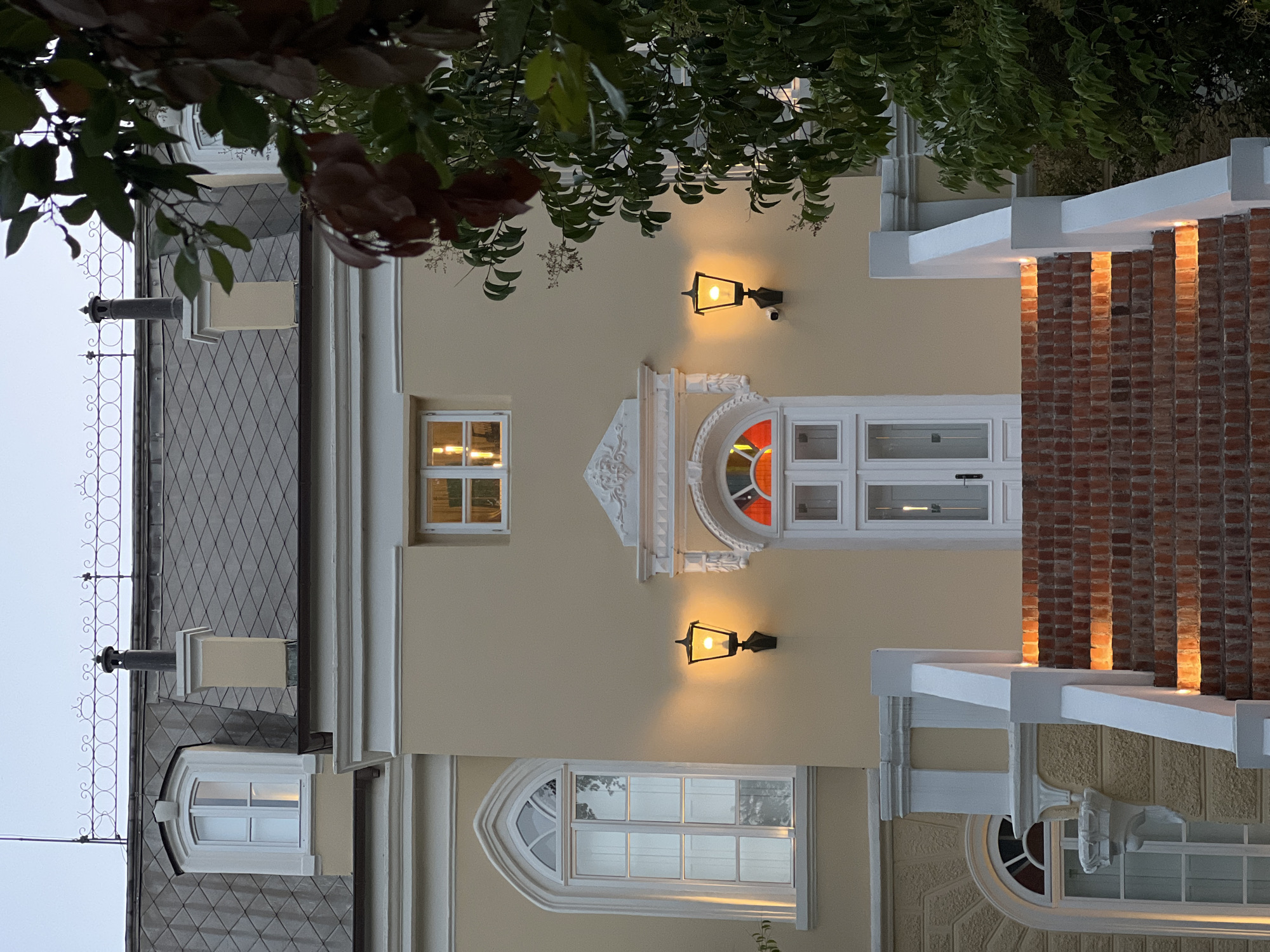 The image shows the entrance of a building, possibly a hotel or a private residence, in Bar, Montenegro. The facade is painted a light yellow, and the roof is gray with a diamond pattern. There are several windows visible, including a large arched window on the left and a smaller rectangular window above the entrance. The entrance itself features a white door with a stained-glass transom window in red, yellow, and blue. Two black outdoor lamps with glowing yellow bulbs are mounted on either side of the door, casting a warm light. A brick staircase with integrated lighting leads up to the entrance, with white railings on either side. Green foliage from a tree partially obscures the right side of the building and the roof. The sky is overcast and appears to be dusk. The scene is devoid of people and any visible activity.