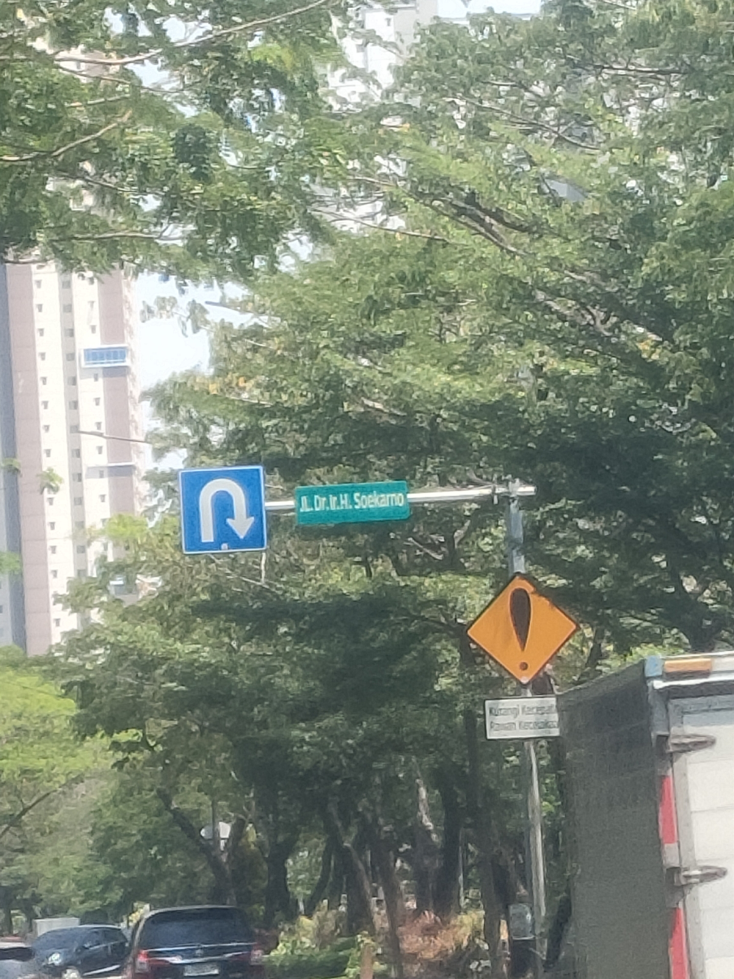 The image depicts a street scene in Surabaya, Indonesia, with a prominent street sign and traffic signs. A blue square sign with a white U-turn arrow pointing down and to the left is visible. Adjacent to it, a green rectangular sign displays the text "Jl. Ir. H. Soekamo" in white lettering. Below these signs, a yellow diamond-shaped warning sign with a black exclamation mark is present. Further down, a smaller white rectangular sign reads "Kurangi Kecepatan" and below it "Rawan Kecelakaan". The background is dominated by lush green trees, with a tall modern building visible on the left. In the foreground, parts of vehicles, including a dark-colored SUV and a truck, are captured. The sky is bright, indicating daytime.