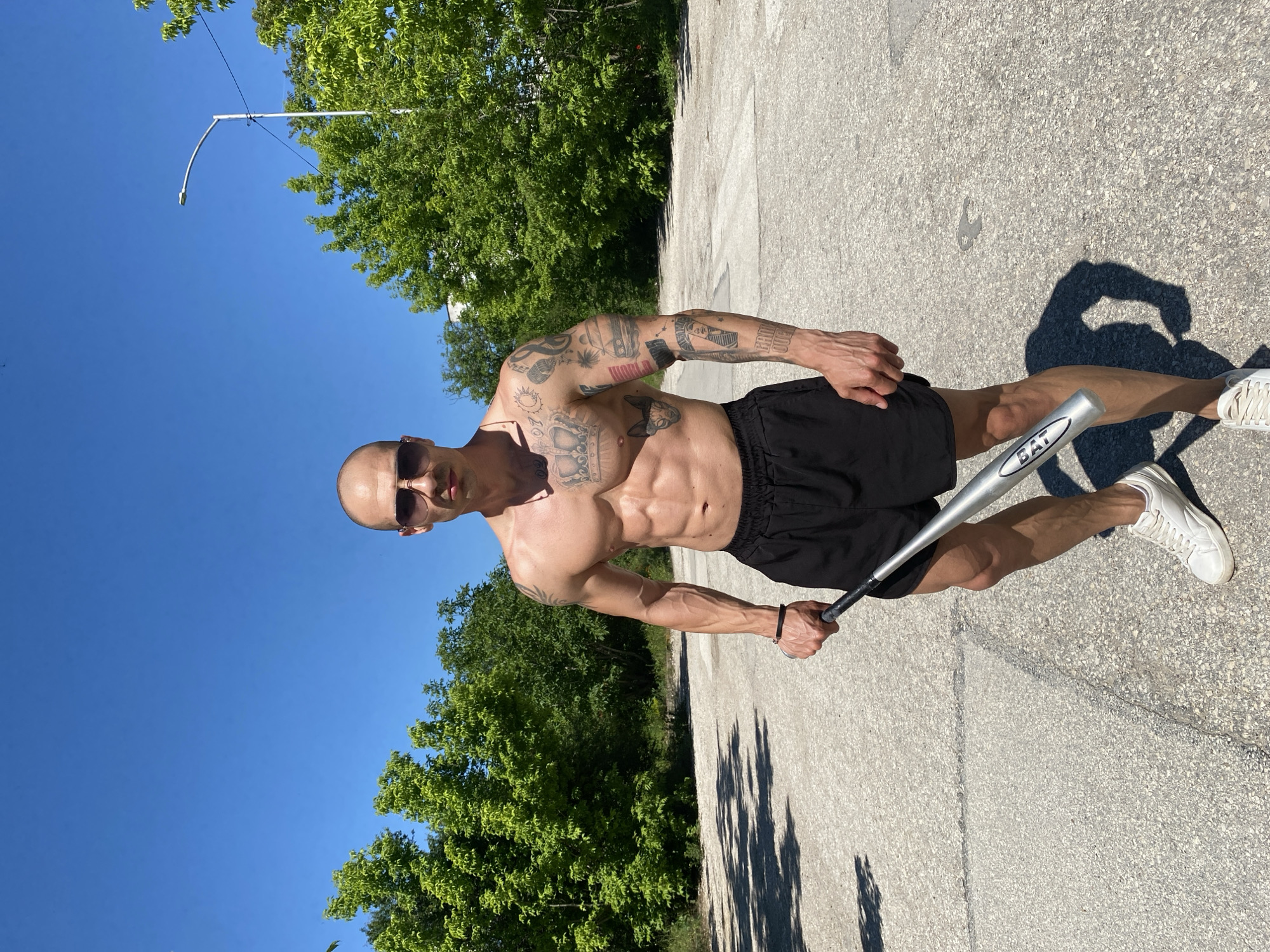 A bald man with sunglasses and tattoos is standing outdoors, shirtless and wearing black shorts and white sneakers. He is holding a silver baseball bat in his right hand. The background includes green trees and a clear blue sky with a streetlight in the distance. The location appears to be a paved area. The location in Pleven, Bulgaria is impossible to be determined from the image provided.