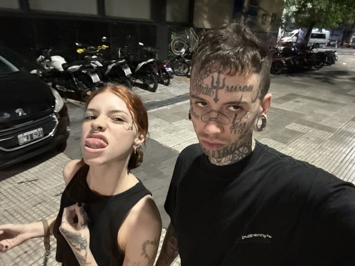 The image captures two young adults, a female and a male, posing for a close-up photograph on an urban street at night. The setting is consistent with Buenos Aires, Argentina, confirmed by the visible Argentine license plate "OMJ 621" on a black Peugeot 208 parked on the left.
The female, positioned on the left, has red hair, a septum piercing, multiple ear piercings, and a black stylized line tattoo extending from her left eye. She wears a black sleeveless crop top, revealing a tattooed forearm and hand. She is sticking her tongue out, looking towards the camera.
The male, on the right, has short dark hair and extensive facial and neck tattoos, including a trident symbol on his forehead, "MAMMA" above his right eyebrow, a bandage-like design on his nose, and "DOOM 1993" on his left cheek. He has large circular ear gauges. He wears a black long-sleeved shirt with "bullbennyt™" embroidered on the chest. He gazes directly at the camera with a neutral expression.
The background features a tiled sidewalk and dark building facades. Numerous motorcycles of various makes are parked along the street, some with visible Argentine license plates. The scene is illuminated by artificial street lighting, indicating evening or nighttime.