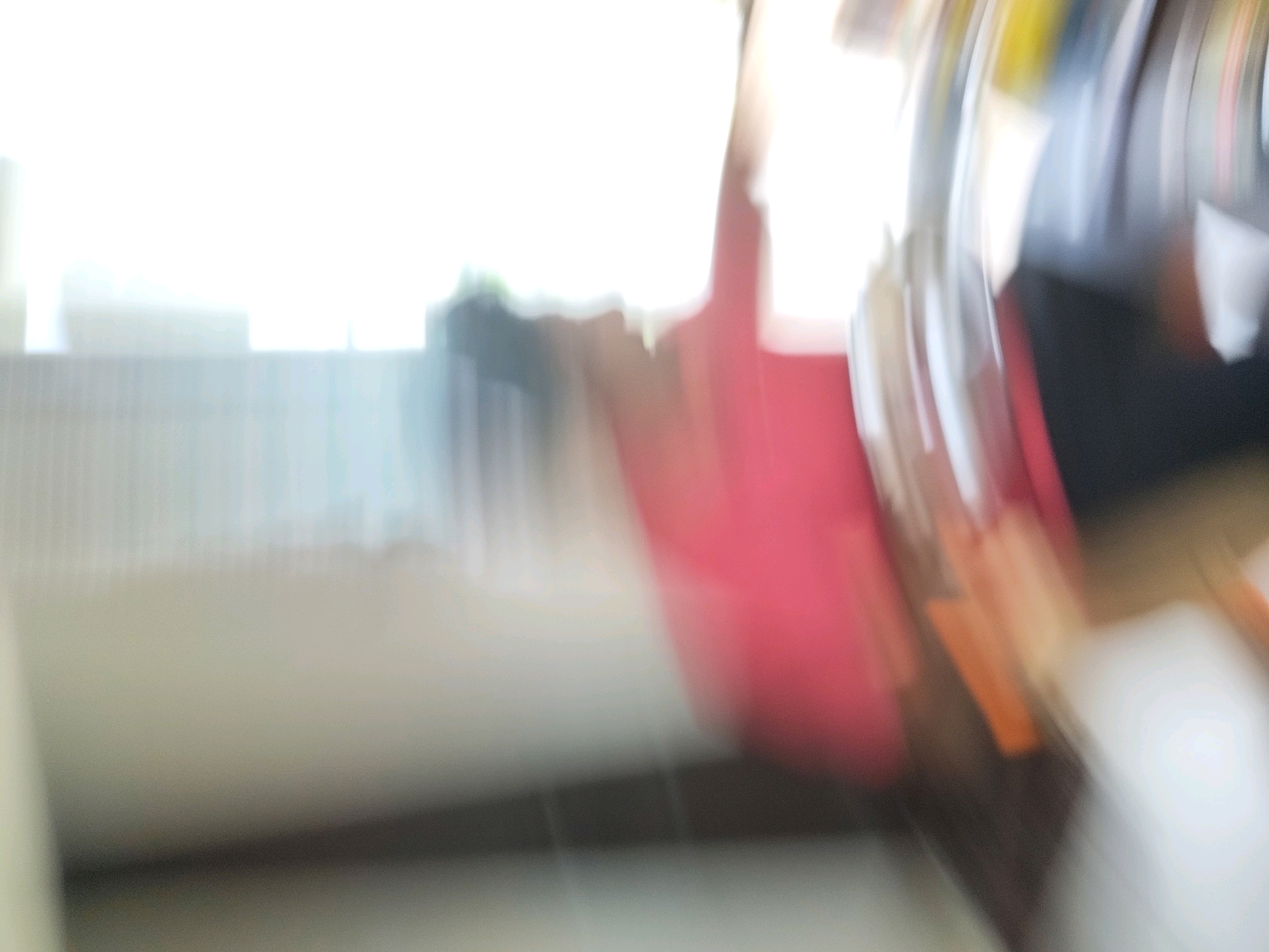The image is an out-of-focus shot, likely taken indoors with natural light coming from a window or bright area on the left. A pink object, possibly a cushion or piece of fabric, is prominently featured in the mid-right of the frame, blurred by motion. To the right of the pink object, there are streaks of color, including yellow and white, suggesting other objects or light reflections. The background is indistinct, with blurred shapes that could be furniture or shelving. The floor in the foreground is also out of focus, showing a textured surface. The overall impression is that of a fast-moving subject or camera, creating a dynamic but unclear scene, likely within a residential or office environment in Tathawade, India.