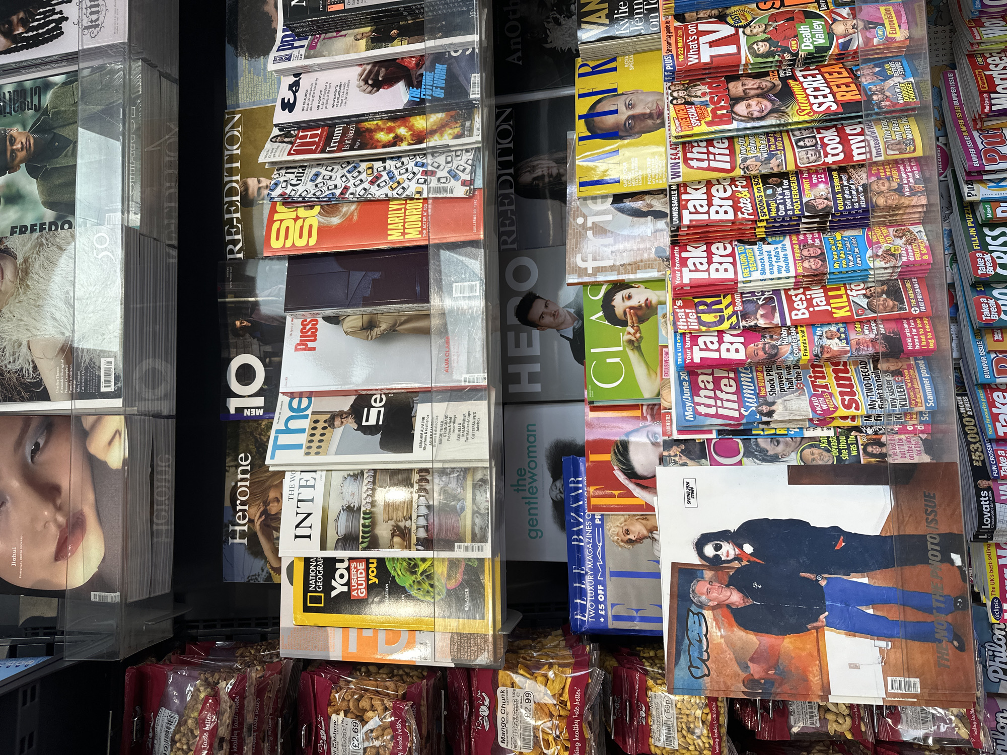The image shows a newsstand display in London, United Kingdom. The shelves are stocked with magazines, including titles like "Elle Bazaar," "Heroine," "Re-Edition," "Take a Break," and "National Geographic." Snack bags of nuts and dried fruit are displayed below the magazines.
