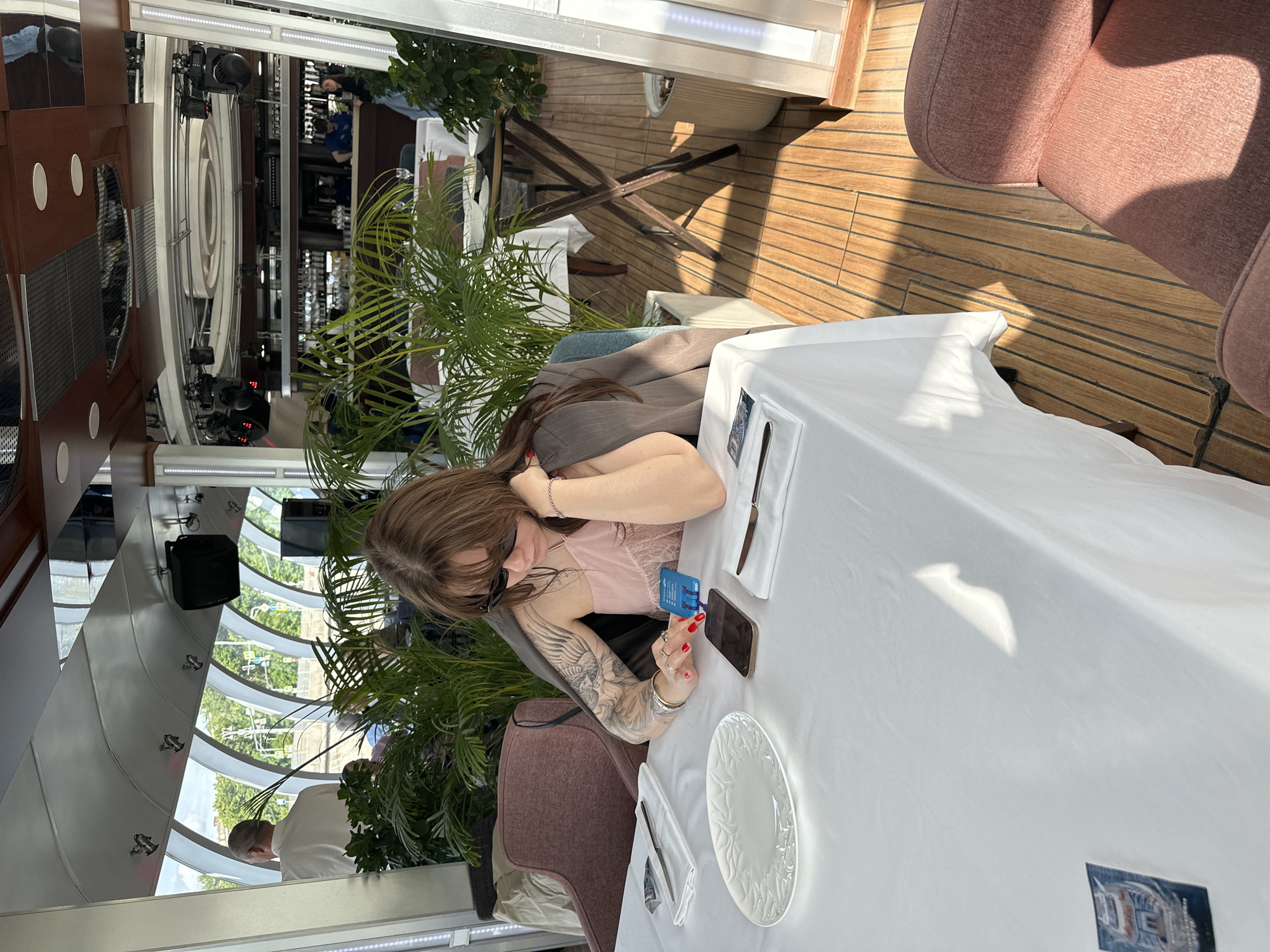 A woman is seated at a table in a restaurant, with a view of a city through curved windows. She is holding a credit card in one hand and a cell phone in the other, potentially engaging in online transactions. The table is covered with a white tablecloth, and a plate, silverware, and napkins are set for a meal. The restaurant appears to have a modern design with indoor plants and possibly a multi-level layout. Based on the window views, the location is likely in Moscow, Russia.