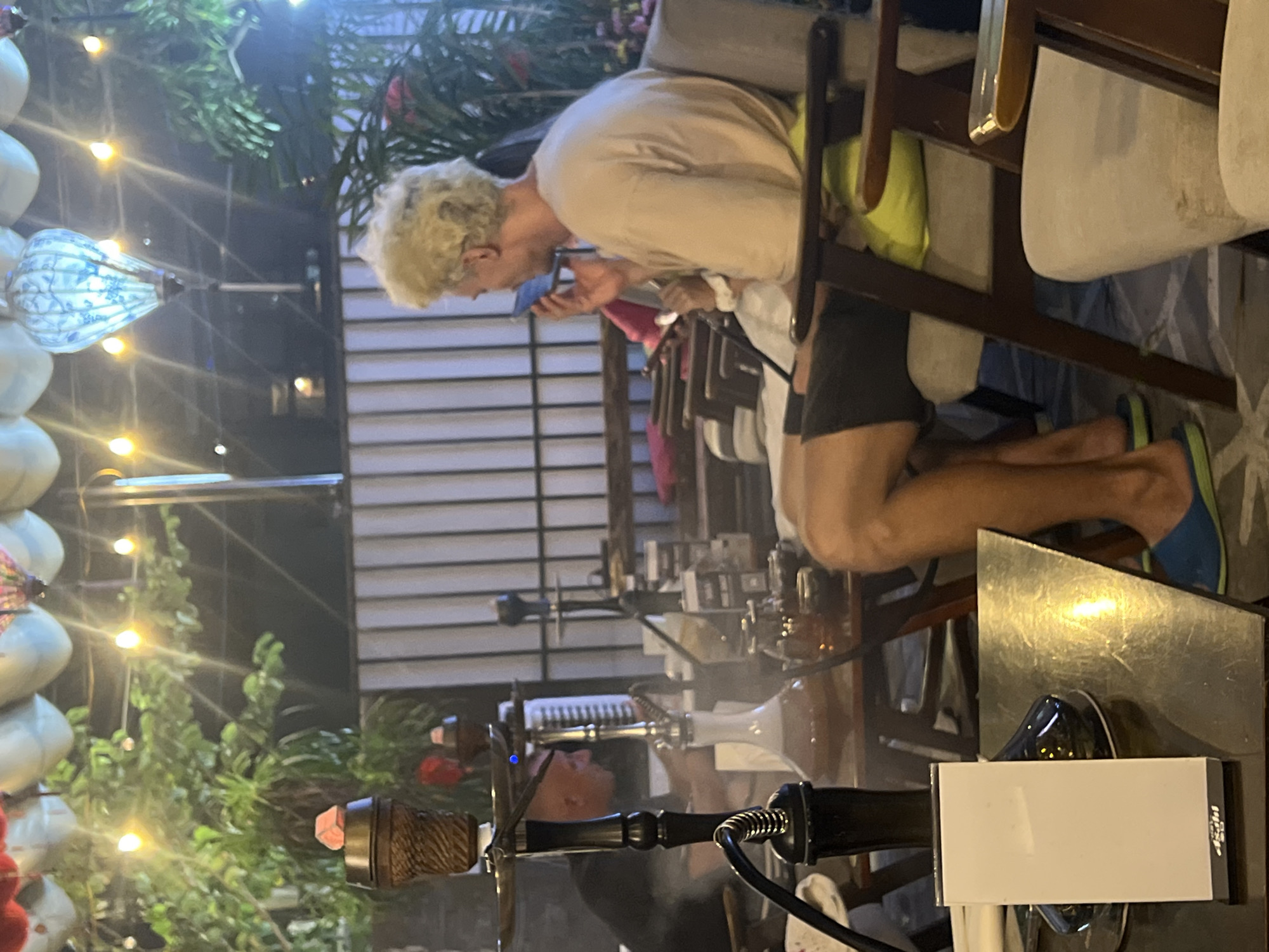 This image captures an evening or nighttime scene at a relaxed cafe or lounge, likely in Đà Nẵng, Vietnam. The setting appears to be semi-outdoor, indicated by the presence of green plants and numerous decorative lights that illuminate the space, creating a warm ambiance.
In the mid-ground, a person with light-colored, curly hair, wearing a light long-sleeved shirt and dark shorts, is seated at a table. This individual is actively engaged with a blue mobile phone held close to their face. They are wearing blue and green flip-flops.
The foreground prominently features a hookah on a table, with visible smoke rising, suggesting it is in use. A part of another person's head is visible behind this hookah, near a second, less distinct hookah setup in the background. On the table in the foreground, next to the hookah, is a white rectangular object with "ASAHI" and some Japanese characters printed on its side. A reflective metallic object is also on the table.
The background reveals more seating, tables, and the structure of the establishment, possibly slatted walls or windows, further emphasizing the open yet intimate atmosphere. The numerous small, warm-toned lights strung across the plants and larger decorative lanterns, including one blue and white patterned light, are the primary light sources, indicating it is dark outside. The overall scene suggests a casual and leisurely social gathering.