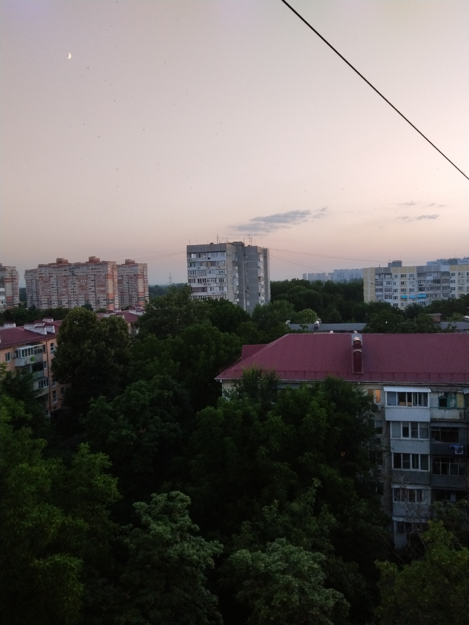The image depicts a cityscape at dusk, featuring multiple residential buildings and a dense canopy of trees. A crescent moon is visible in the pale sky, along with a thin wire stretched diagonally across the upper right portion of the frame. The buildings are typical of Soviet-era architecture, with varying heights and facades. In the foreground, a building with a red tiled roof and balconies is partially obscured by dark green trees. The sky transitions from a light pinkish hue near the horizon to a more muted gray at the top. There are no people or discernible actions captured in the scene. The overall impression is of a residential area in a Russian city during twilight.