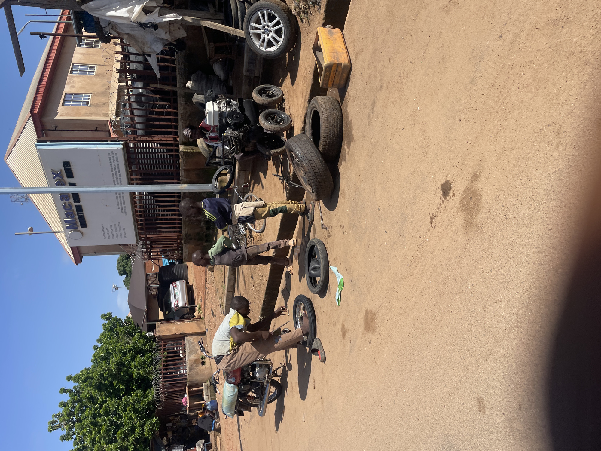 In Gura Top, Nigeria, on a sunlit day, a man wearing a yellow shirt and brown pants is working on a motorcycle, which is propped up by its kickstand. He is bent over, appearing to fix a tire, with another tire and a piece of green cloth on the ground nearby. To his right, a young boy in a green and gray outfit stands with his arms crossed, watching the activity. Further back, a man wearing a red vest and a cap is seated next to a four-wheeled engine. The scene is set on a dirt road with a concrete curb. In the background, a building with a sign that reads "Maca ex MARKETING COMPANY LIMITED" is visible, along with trees and a fence. Several other tires are scattered around the area, some of which are stacked.