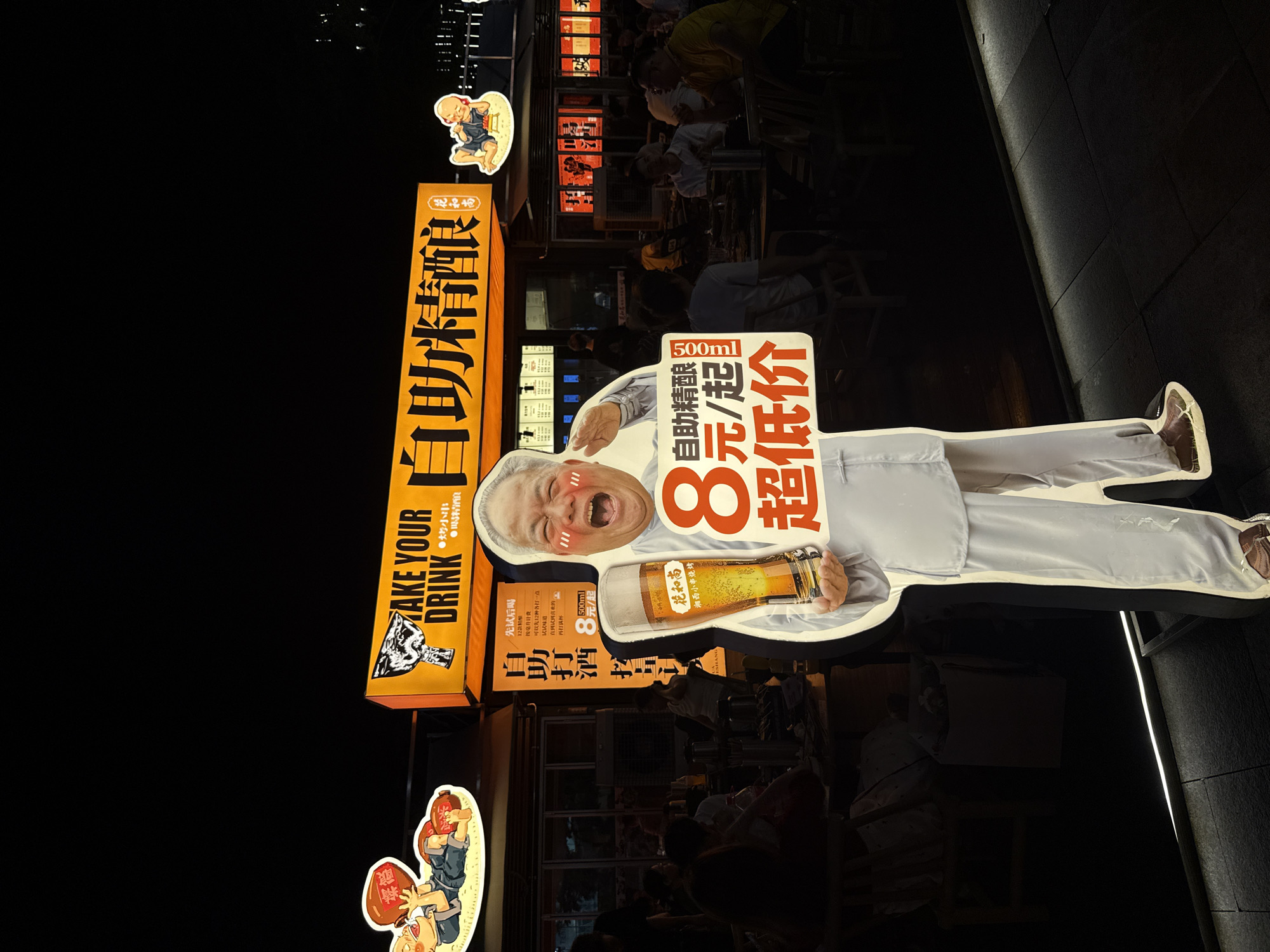 The image depicts a nighttime scene on the sidewalk in Haizhu District, China. A life-sized, cutout advertisement of an elderly man wearing traditional Chinese clothing and enthusiastically holding a beer glass stands on the right. The advertisement promotes "take your drink", a beer named "Heshang" from Xiangxi BBQ skewers, and is written in both English and Chinese, advertising 500ml of the beer for ¥8. Other signs with Chinese characters are also displayed in the background of the scene. In the dim background, people are seated at tables and chairs, seemingly patrons of the establishment the advertisement is for. The floor has dark tiles and the environment is dimly lit.