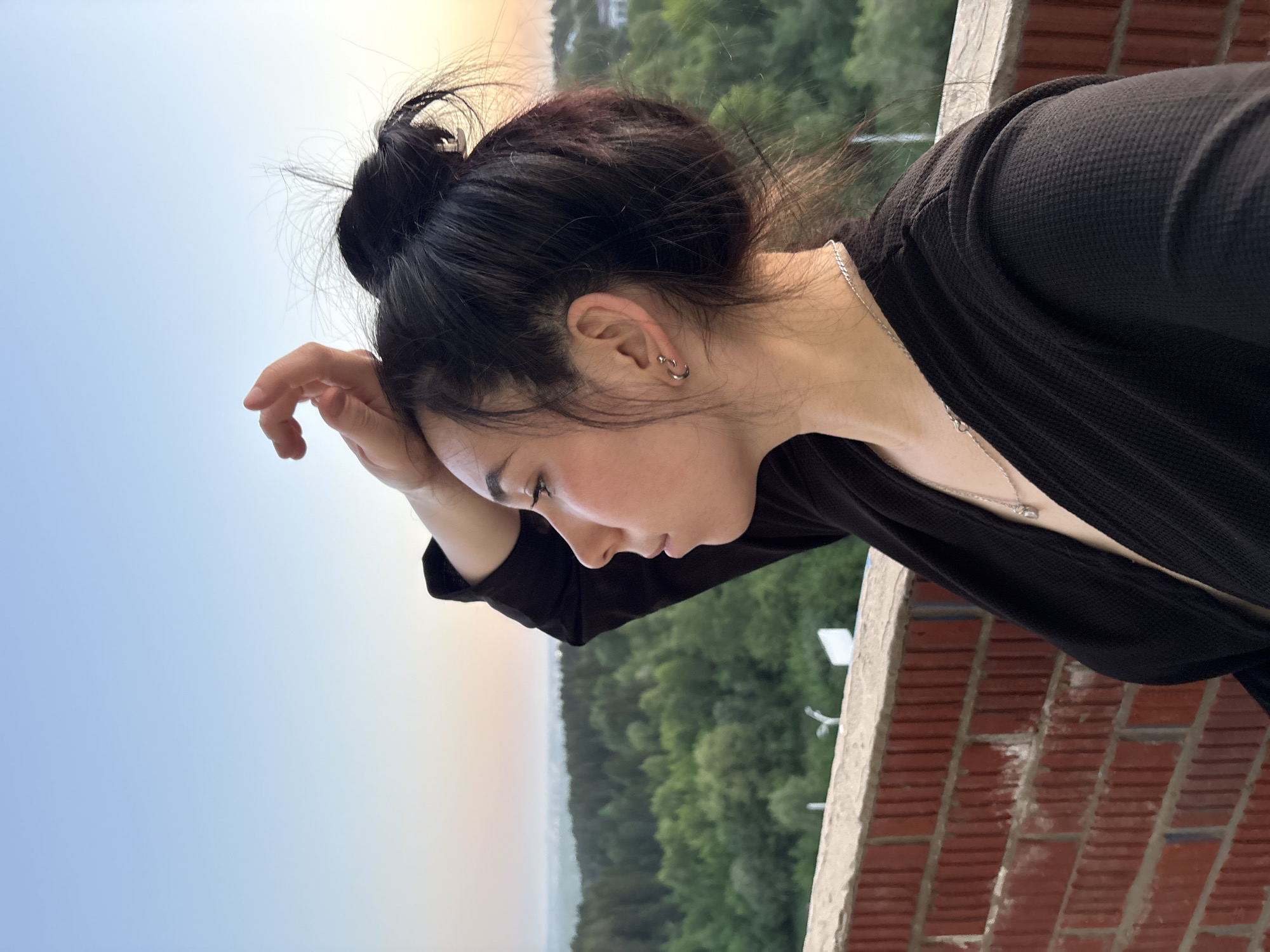 A sideways, close-up shot features an East Asian woman with her dark hair in a messy bun. She wears small hoop earrings and a delicate silver necklace. Her eyes are closed, and her hand rests on her forehead. She is wearing a black, textured, long-sleeved garment.
The background reveals a blurred vista, likely a cityscape blended with greenery. A brick and concrete barrier or railing is visible at the edge of the frame. The setting could be Izhevsk, Russia, given the user's prompt, however there is no obvious visual confirmation of the location within the image.