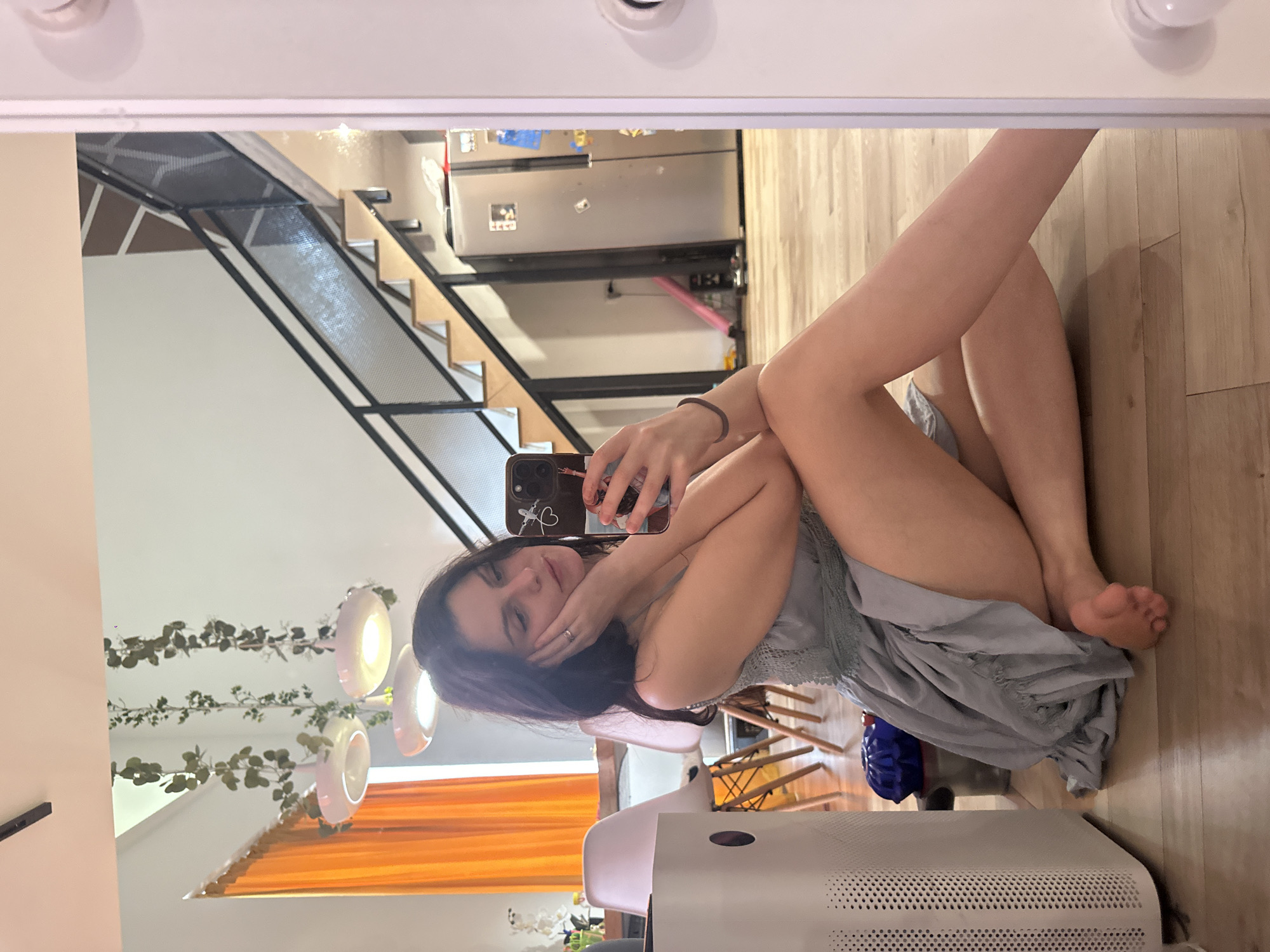 A young woman with dark hair is sitting on a wooden floor, taking a mirror selfie with her phone. She is wearing a light blue dress and is resting her head on her hand. In the background, a staircase with a black railing is visible, as well as a refrigerator and orange curtains. To the left, a white air purifier is present. The scene appears to be indoors, likely in a modern apartment or house.