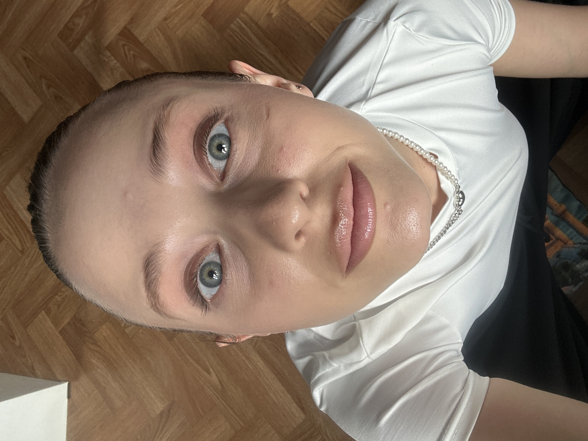 The media file is a close-up, overhead shot of a young person, likely female, lying down on a wooden floor. The subject has fair skin, light-colored eyes, and light brown hair pulled back. She is wearing a white t-shirt with a pearl necklace. Her lips are glossy, and she appears to be wearing makeup, including mascara and possibly a subtle blush. The lighting suggests it is daytime, with natural light illuminating the scene.
The subject's gaze is directed upwards towards the camera, with a neutral to slightly curious expression. The wooden floor is visible in the background, showing a parquet pattern. The composition is a tight crop, focusing primarily on the subject's face and upper torso. There are no other people or objects clearly visible that provide additional context about the activity or event. The overall impression is one of a casual, personal moment captured from an unusual perspective. The setting appears to be indoors, given the floor and the quality of light. There is no visible text in the media file.