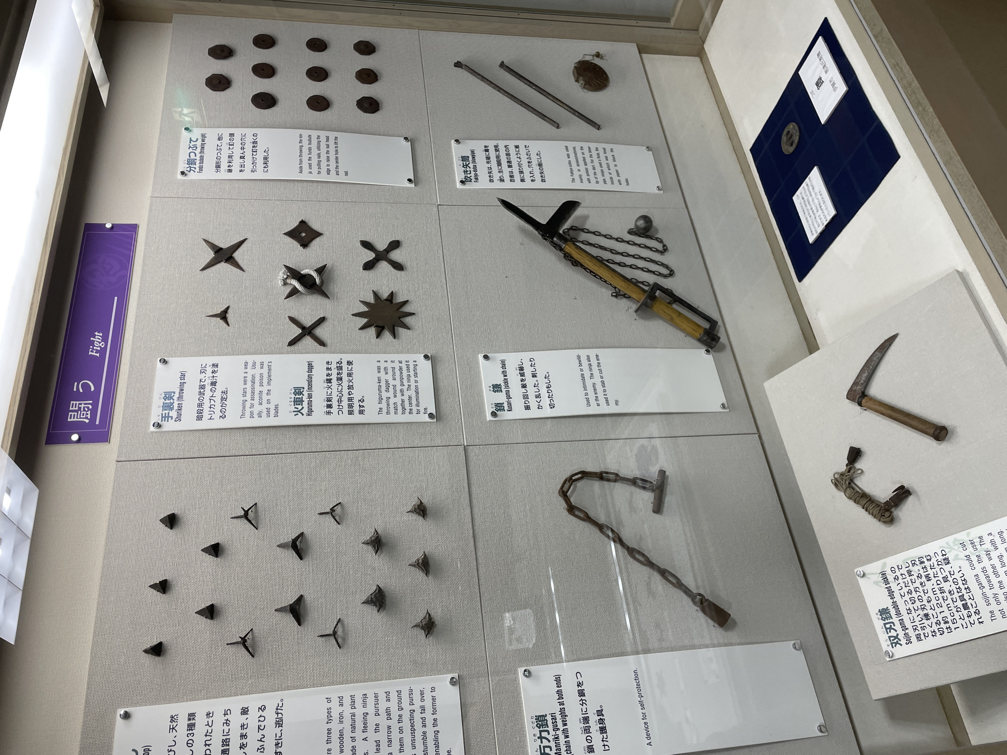 The image shows an exhibit of ninja weapons in the Iga Ninja Museum, located in Iga City, Japan. The display features various historical artifacts, including shuriken (throwing stars) of different designs, a higuruma-ken (incendiary dagger), a funso-tsubute (throwing weight), a kusari-gama (sickle with chain), and a fukiya-duto (blowpipe). The weapons are arranged in a clean, museum setting with informational text panels in Japanese and English describing each item. The overall environment is well-lit and organized, highlighting the historical significance of the ninja artifacts.