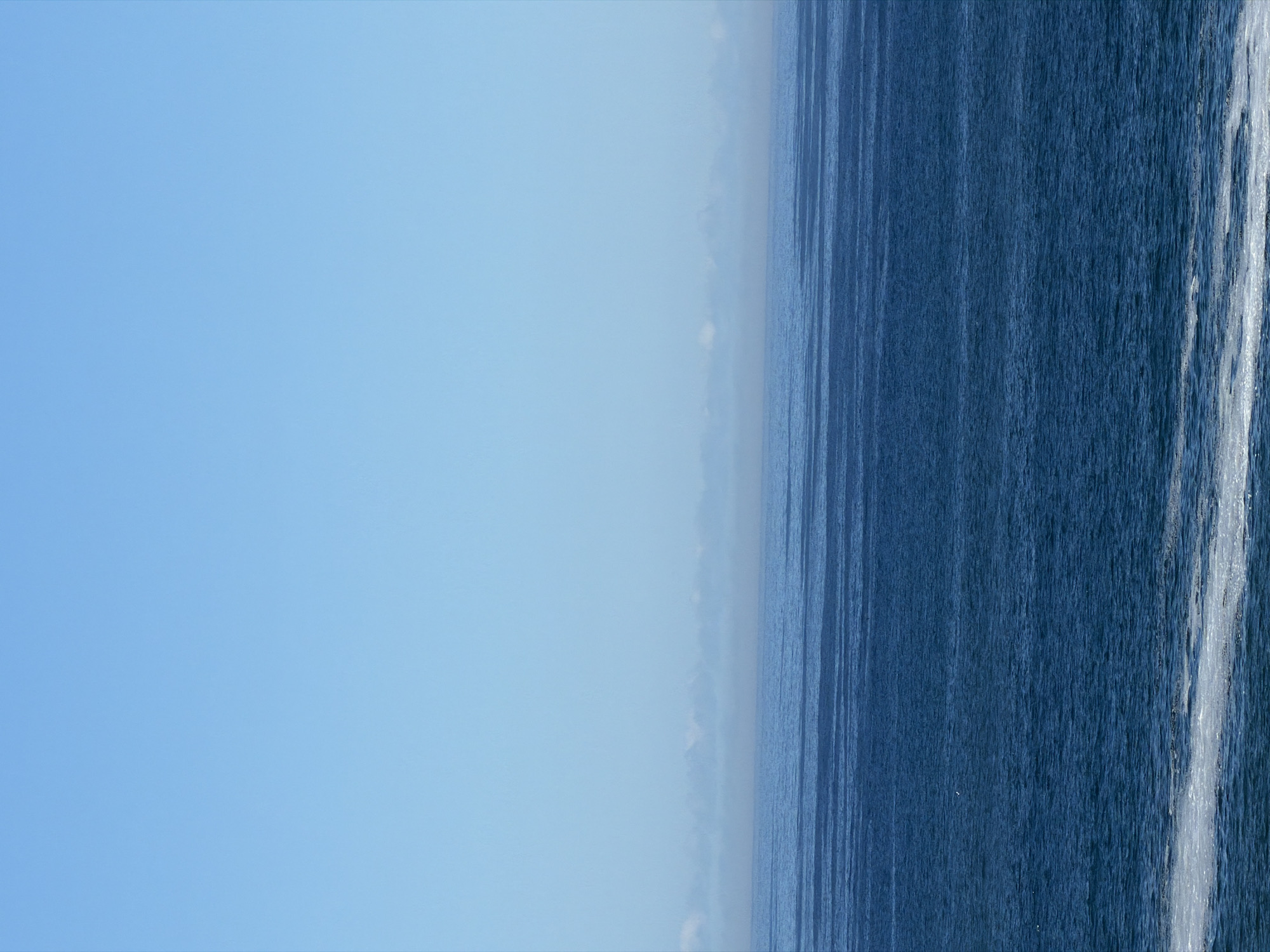 The image depicts the ocean meeting the sky, likely near Cape Town, South Africa. The left side of the image shows a pale blue sky, transitioning to a hazy horizon line. The right side displays the ocean with visible water texture and some white foam near the right edge. The image has been rotated, so the sky appears on the left and the ocean on the right. There are no people or discernible objects present.