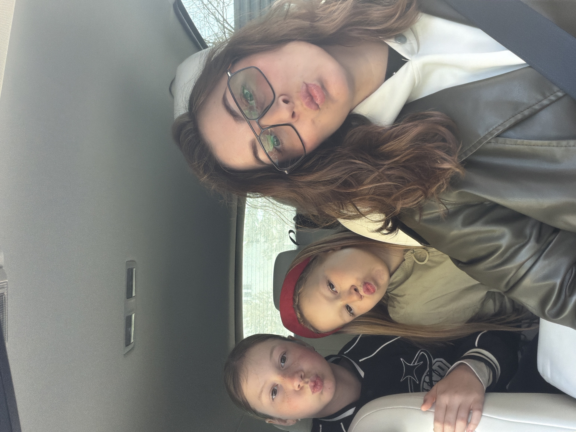 Three individuals are captured in what appears to be the interior of a vehicle. The setting is suggested by the interior components visible, such as seats and panels.
An adult with glasses is positioned at the top of the frame. Below, there are two children. All three are making a kissing face.
There is no information in the image to suggest the specific location as Dubovaya, Russia.