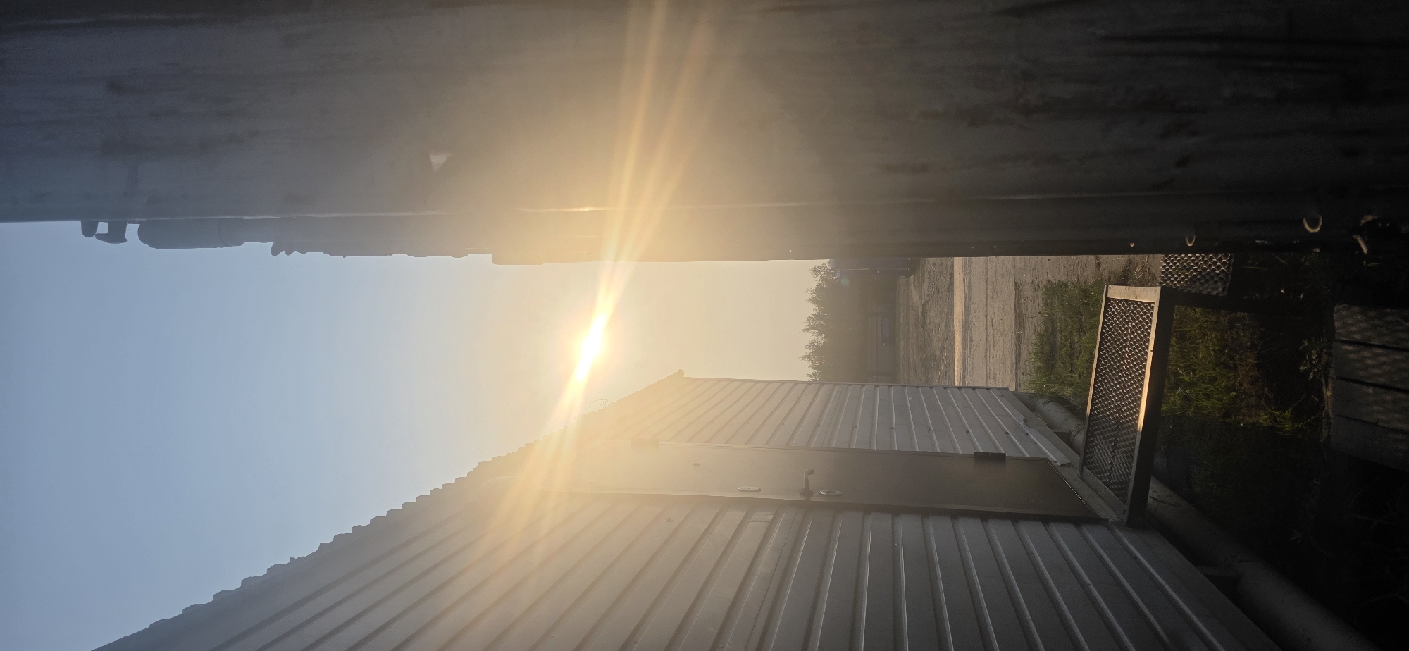 This image depicts an outdoor scene with a bright sun low in the sky, suggesting either sunrise or sunset. The camera is oriented sideways, with the top of the frame showing a dark, metallic structure, possibly the underside of a larger object or a ceiling. Below this, a wide expanse of clear sky dominates, with the sun emitting strong rays and creating a lens flare effect.
In the lower half of the image, a corrugated metal building with a door is visible. The building's metallic surface reflects the bright sunlight. To the right of this building, there is an area with grass and some sort of mesh-like object, possibly a grate or a screen. Further in the background, a dirt or gravel path is visible, bordered by trees and vegetation. The overall setting appears to be industrial or rural, with structures and open space.
The time of day is clearly indicated by the low angle of the sun and the warm, golden light, characteristic of early morning or late evening. The weather appears clear, with no clouds visible in the sky. The location context of Nizhnevartovsk, Russia, implies a possible northern setting, and the presence of what appears to be a simple, utilitarian building further supports a functional or industrial area. No people are visible in the frame. There is no visible text in the image.