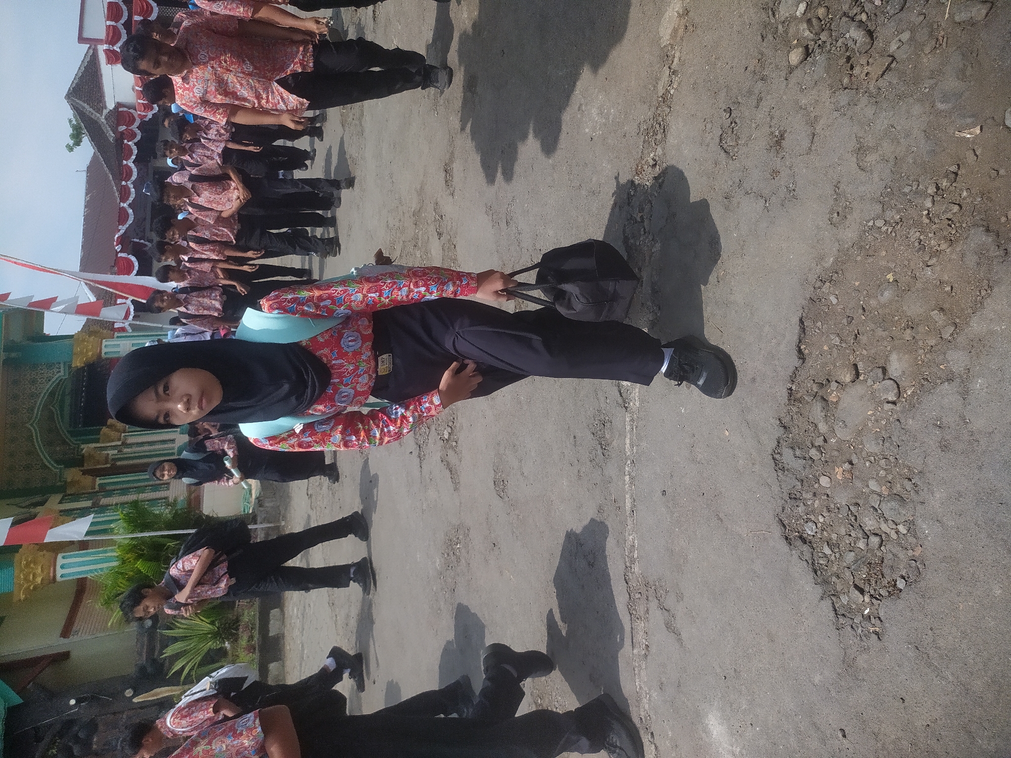 The image depicts a group of students walking on a paved ground in Candirejo, Indonesia. The students are wearing school uniforms which consist of patterned shirts and black pants. One student in the foreground, a girl with a black hijab, is carrying a black tote bag in her right hand and a light blue backpack on her left shoulder. She is looking towards the camera and walking forward. Other students are walking in the background, some with backpacks. The ground is a mix of asphalt and dirt with some potholes. In the background, there is a building with a green and gold facade, and a red and white flag. The sky is overcast.