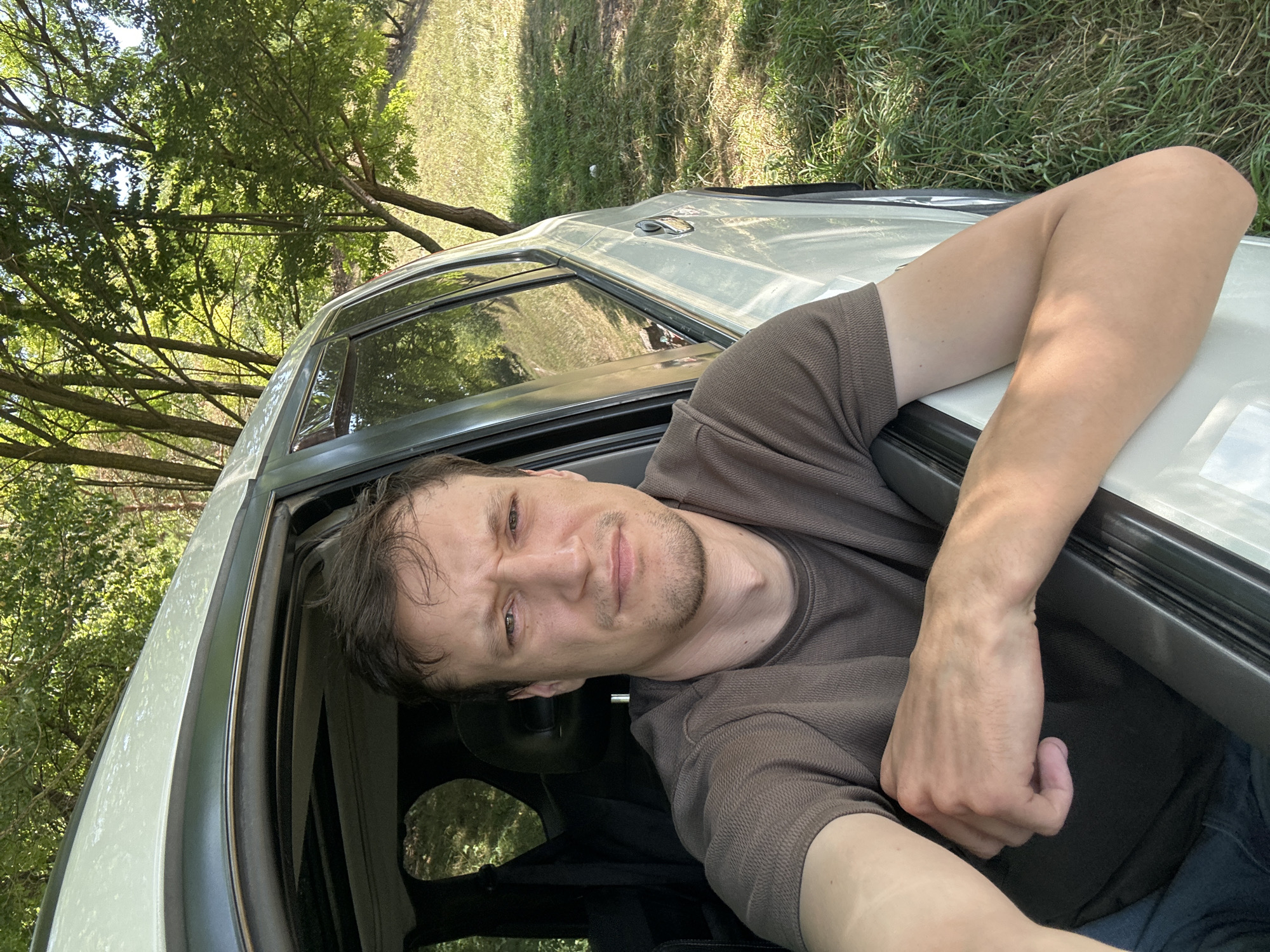 A medium shot selfie is captured from inside a vehicle, looking out through an open door. A man with short brown hair and a scruffy beard is positioned in the driver's seat, leaning his arm on the open door frame. He is wearing a brown t-shirt. The vehicle's interior is partially visible, with a black seat and a sunroof. Outside, the scene is rural, with lush green grass, trees, and sunlight filtering through the leaves, suggesting an outdoor, natural setting. The location is identified as Makiv, Ukraine.