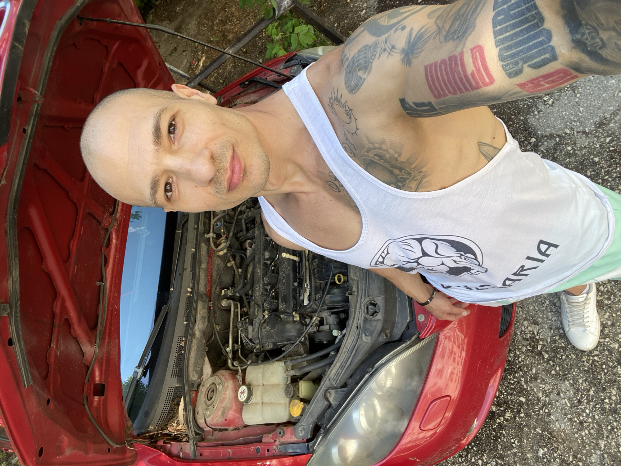 A bald man wearing a white tank top and light green shorts is standing in front of an open red car hood. The car's engine is visible. The man has tattoos on his left arm and a bracelet on his left wrist. The background consists of foliage, a metal fence, and a paved surface. The tank top has "BULGARIA" written on it. There is no information present in the image that can specifically confirm the location is in the city of Pleven.