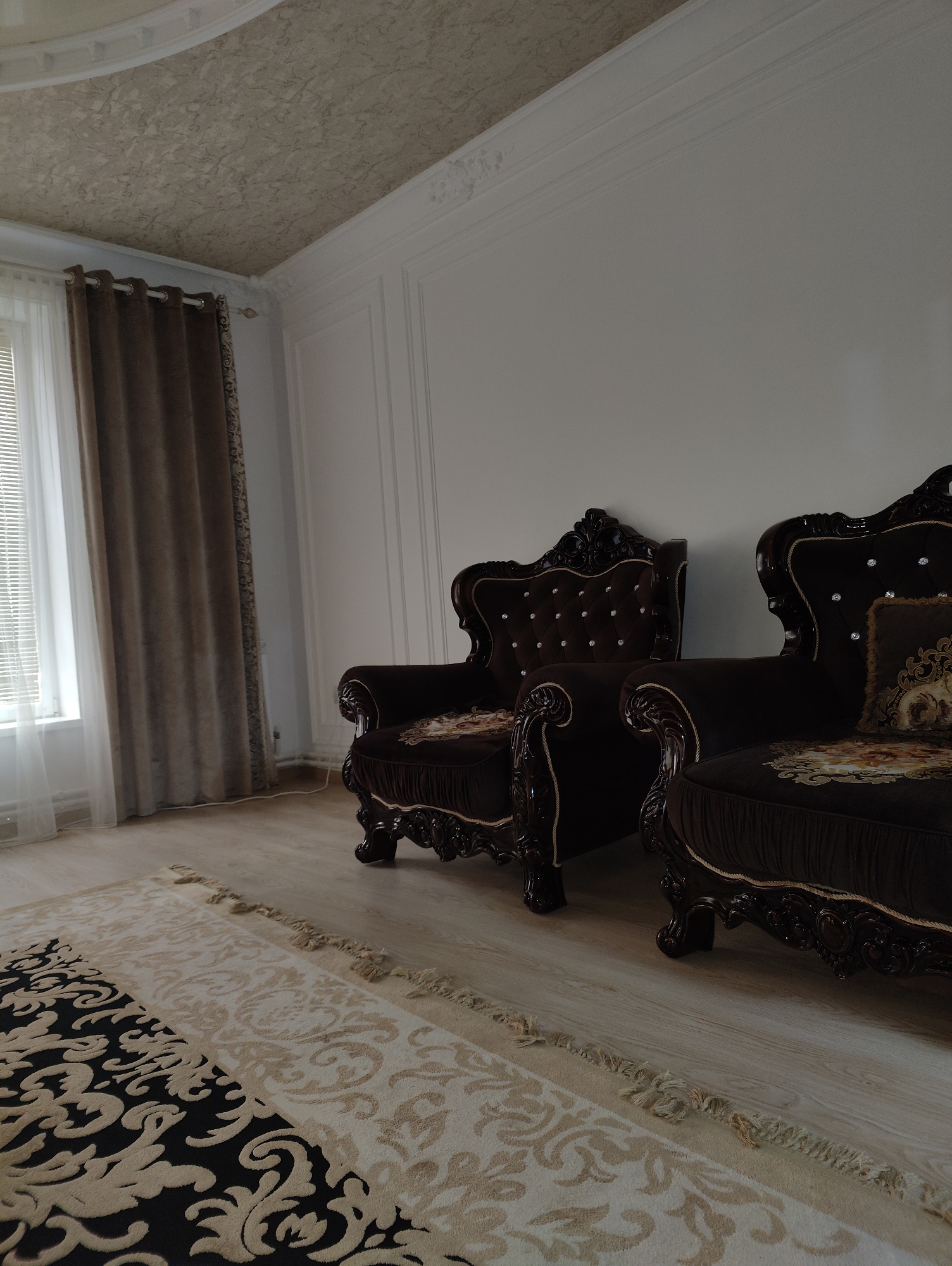 The image depicts an interior scene in a room. Two ornate, dark brown velvet armchairs with tufted backs and crystal accents are prominently featured. The armchairs have carved dark wood frames and decorative trim. One armchair has a decorative throw pillow with a gold and cream floral pattern. In the foreground, a large rug with a black and beige damask pattern covers the wooden floor. A large window with sheer white curtains and heavy brown drapes is visible on the left side of the frame. The ceiling has a textured surface and ornate molding. The walls are painted white and feature decorative molding. The location is stated to be in Nuradilovo, Russia, though no specific indicators of this location are visible within the image itself. There are no people or animals present in the image.