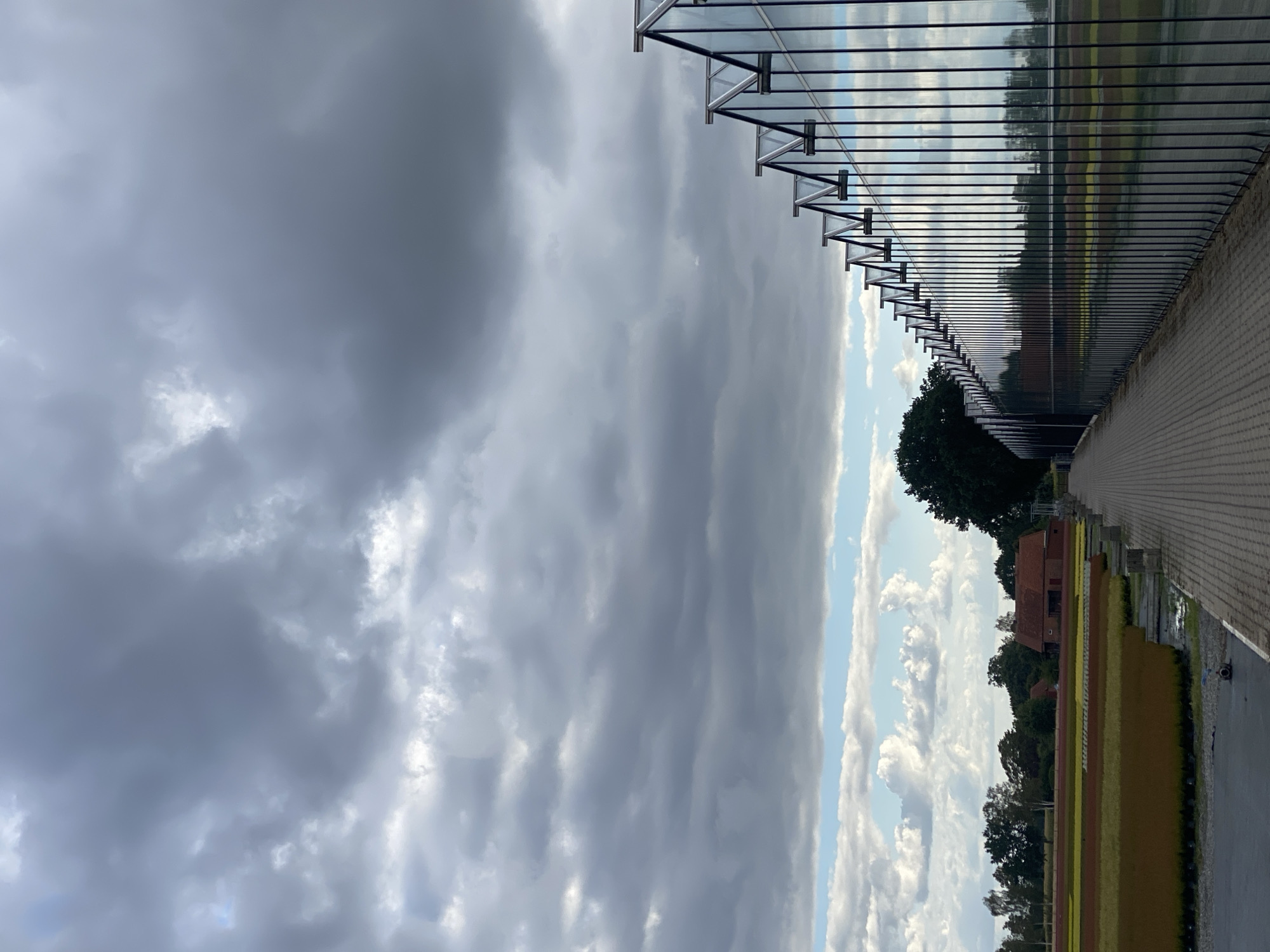 The image shows a modern greenhouse structure with a glass facade on the right side, reflecting the sky and surrounding landscape. A paved pathway runs alongside the greenhouse. In the background, there is a field with rows of colorful plants, a traditional brick house, and trees under a cloudy sky. The scene is set in the rural outskirts of Schwarme, Germany, a municipality in the district of Diepholz, Lower Saxony. The overall impression is one of agricultural industry meeting modern architecture in a natural setting.