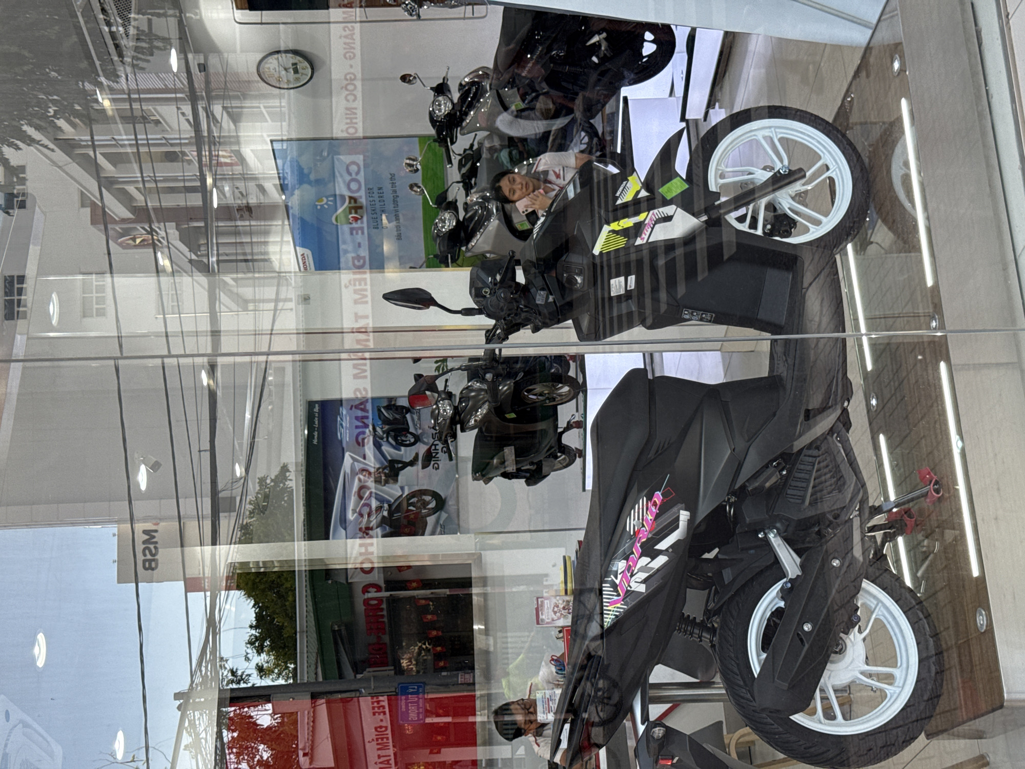 The image depicts the interior of a motorcycle dealership, viewed through a glass storefront. In the foreground, two black scooters are prominently displayed. The scooter on the left has "VARIO" text on its side. The scooter on the right has "HONDA" branding. Behind these, several other scooters are visible, some silver and some black.
Inside the dealership, two individuals are present. On the left, a person wearing glasses and a white shirt is seated and looking down, possibly at paperwork or a device. On the right, a woman wearing a white shirt with a red collar is standing and holding a pink mobile phone to her ear. The background shows reflections of buildings and trees, indicating an urban setting. Signs are visible in Vietnamese, with some text appearing to be reversed due to reflection. One sign reads "MSB" with reversed text below it, and another appears to be a Honda advertisement with text related to "coffee" and "children". The overall environment suggests a commercial establishment in a city. The presence of Vietnamese text and the style of the buildings visible in the reflections strongly suggest the location is in Vietnam. The specific mention of Nha Trang, Vietnam in the prompt implies this is the intended location context for the image.
