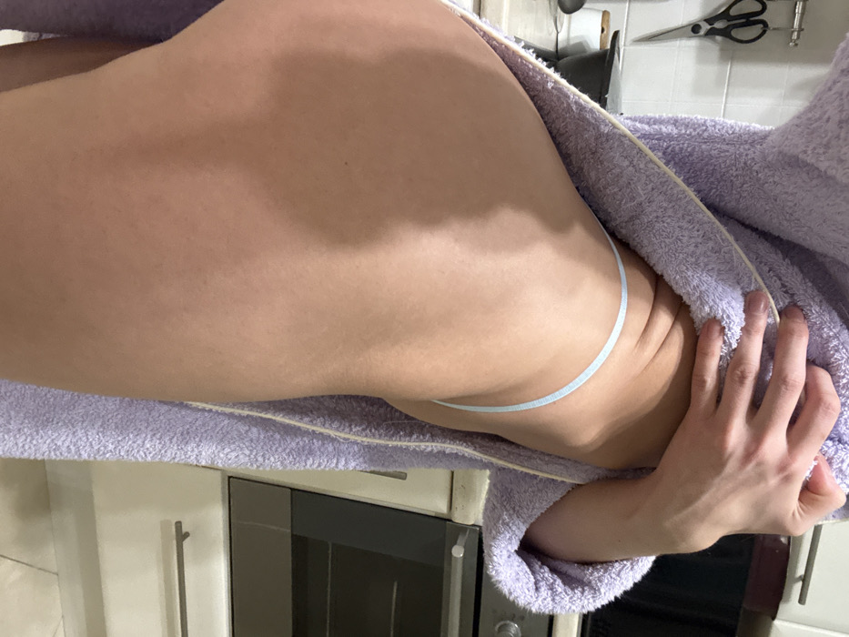 The image, rotated 90 degrees counter-clockwise, presents a close-up view of a person's lower torso and gluteal region. The subject, likely female, is lying on their side, partially covered by a light purple, fluffy bathrobe which is draped open. They are wearing light blue string thong underwear, visible along their hip. One hand is seen resting on the bathrobe.
The background indicates an indoor domestic setting, specifically a kitchen or utility area. Light-colored cabinets with metallic handles are visible. Below the subject's body, an appliance resembling a microwave or oven with a dark glass door is present. Hanging on the wall in the upper background are a pair of black scissors and other indistinct kitchen implements. The lighting appears to be even, typical of an indoor environment. The activity depicted is a static pose. The scene takes place in a home, with the provided context placing it in Saint Petersburg, Russia. There is no visible text in the image.